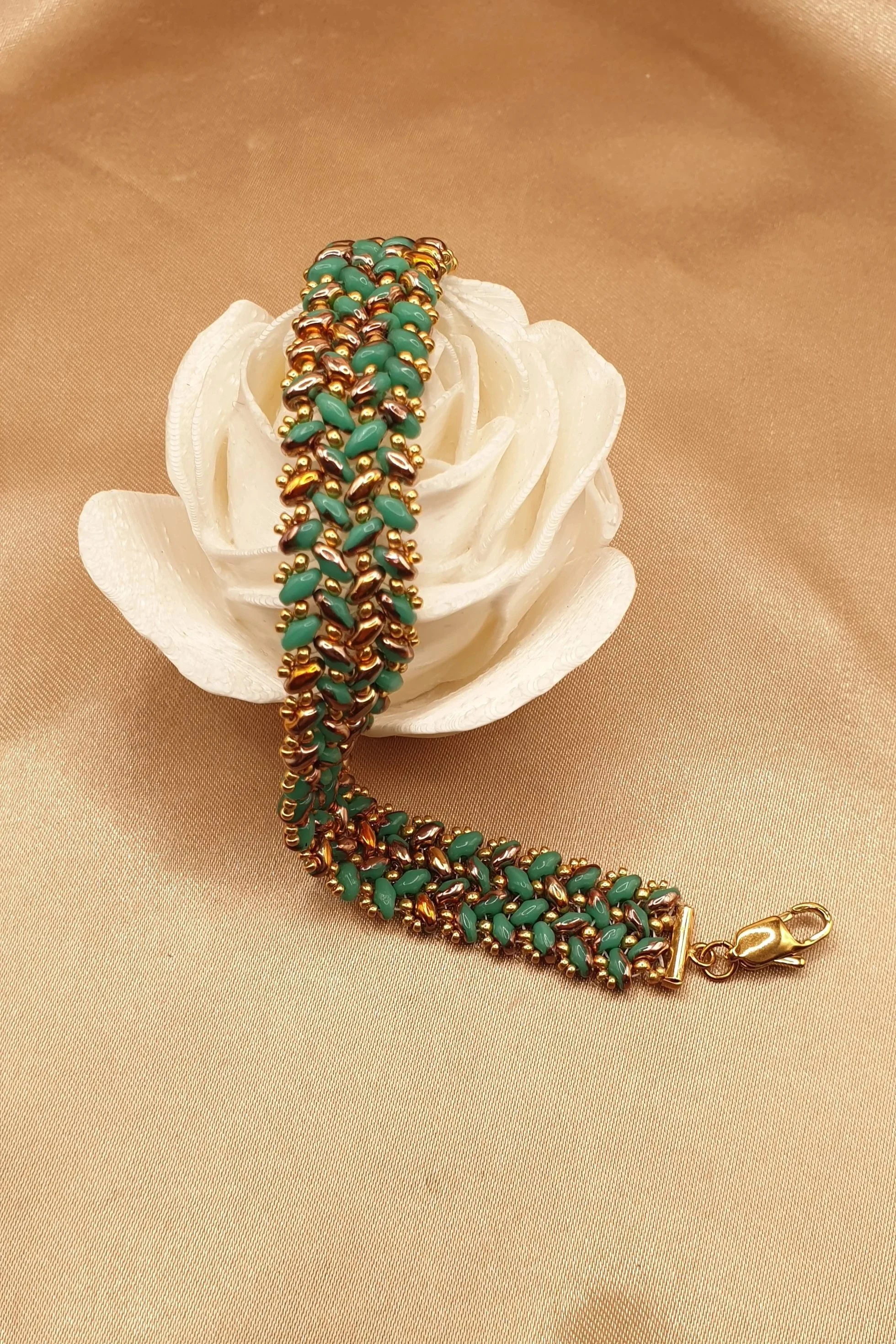 Bracelet Oasis d’Or en perles vert turquoise et dorées, disposé en courbe devant une fleur blanche