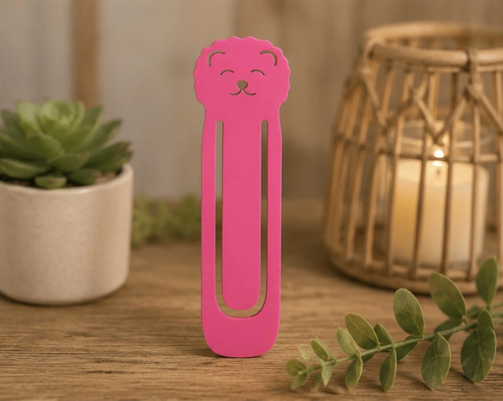 Pink bookmark with a cute lion face design, standing upright on a wooden surface near potted plants and a lantern with a candle inside.