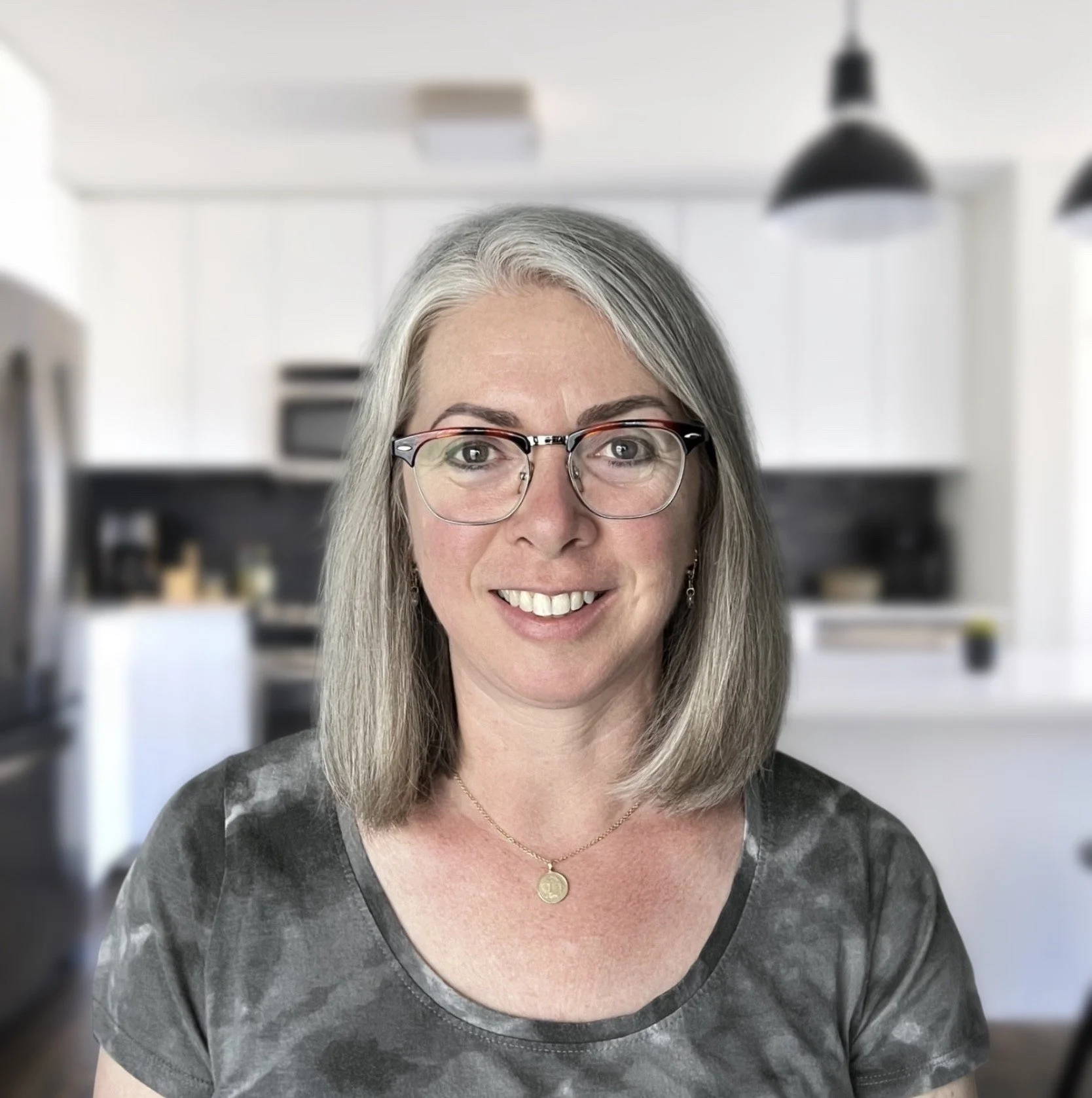 Portrait of a middle-aged woman with silver hair, glasses, and a friendly smile, in a modern kitchen with black and white decor.