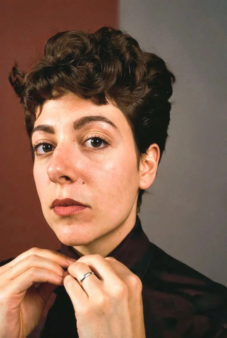 Close-up portrait of a young person with short, dark, curly hair, wearing a dark top, doing up the top button of their shirt, against a dark background.
