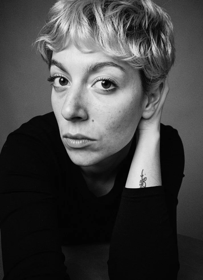 Black and white photo of a woman with short, wavy hair, looking directly at the camera with a serious expression. She is resting her head on her right hand, with a tattoo visible on her wrist.