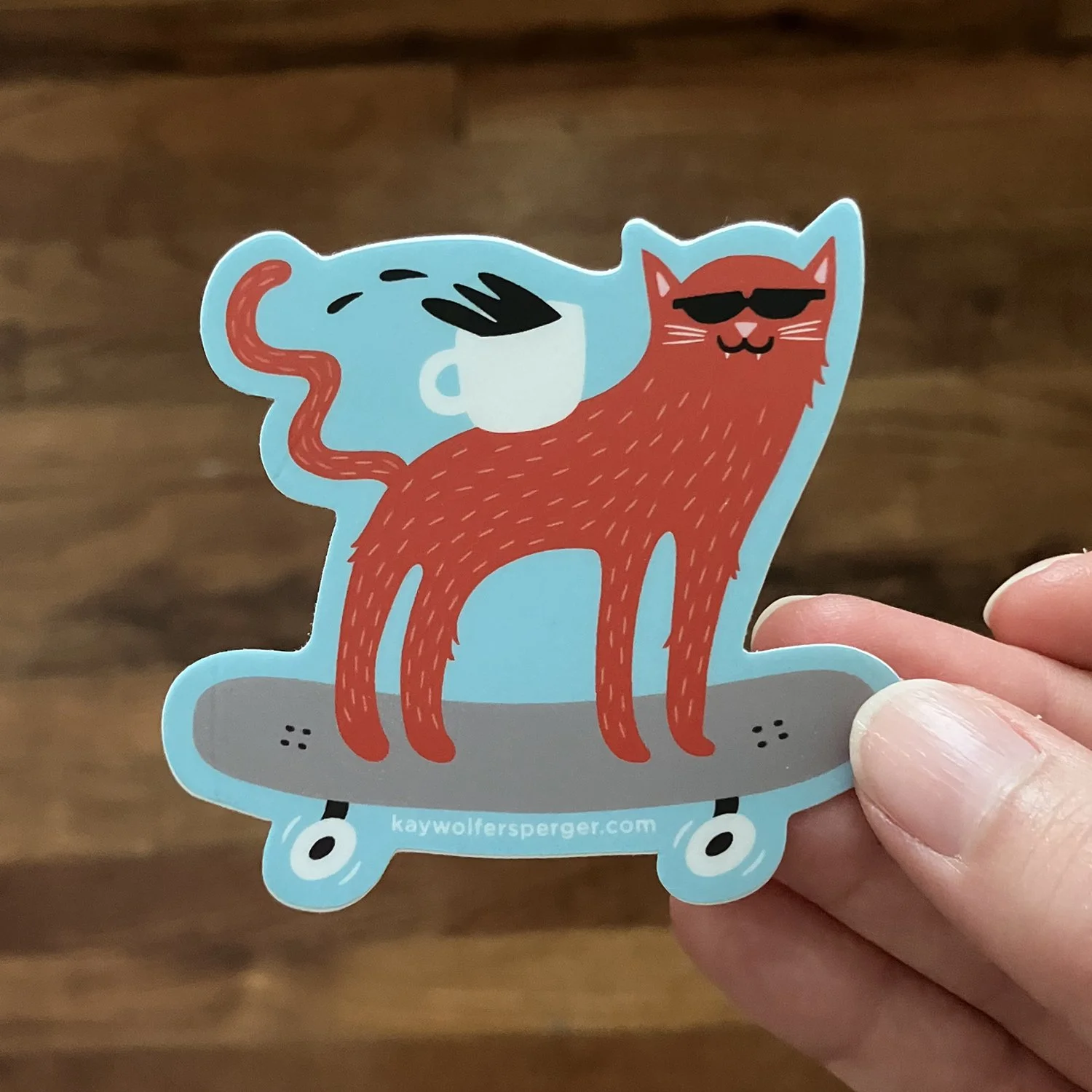 A sticker of a red cat wearing sunglasses, standing on a skateboard with a blue coffee cup on its back, all drawn in a playful cartoon style.