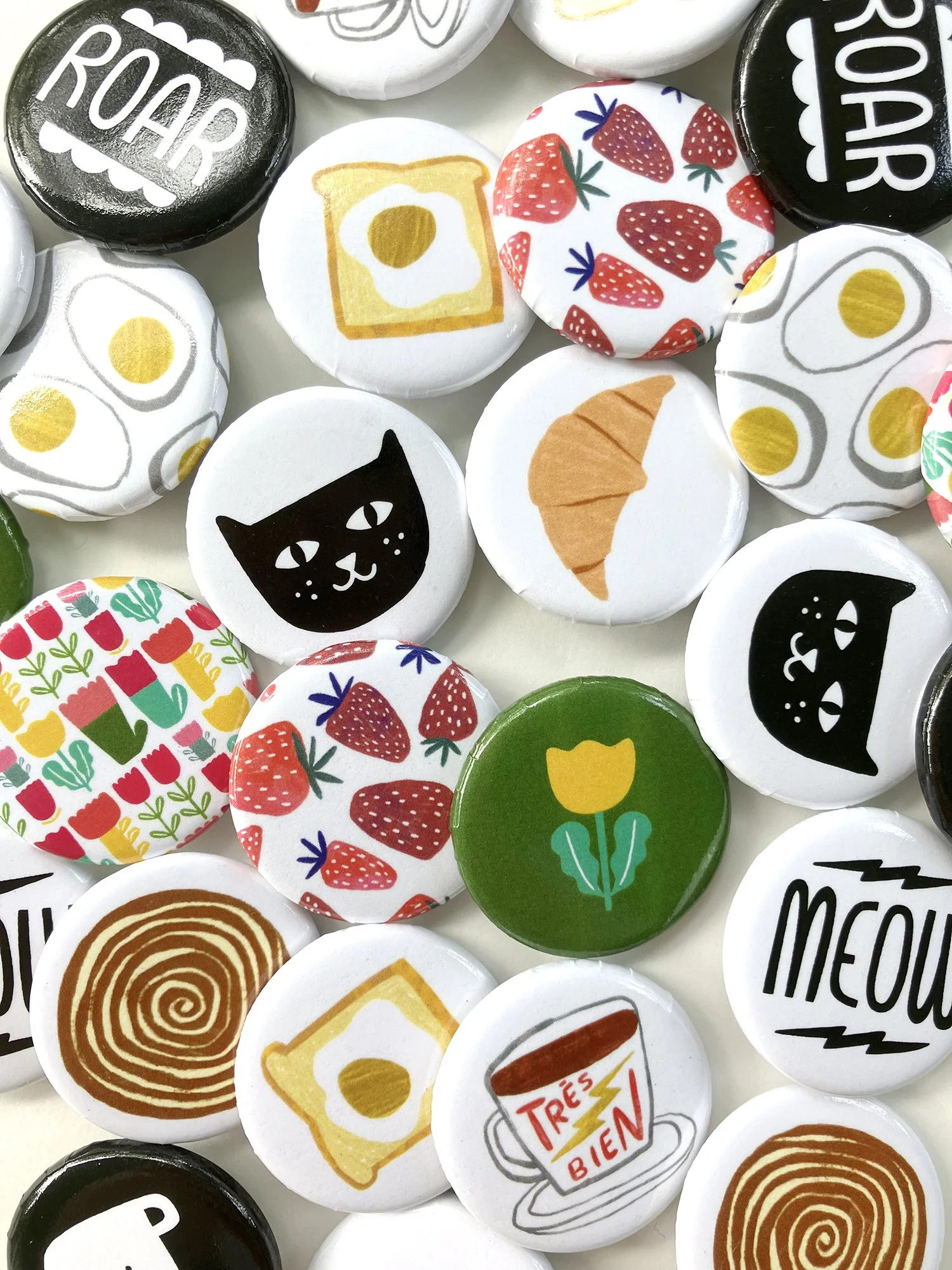 Collection of colorful buttons with various food and drink illustrations, including strawberries, fried eggs, croissants, a cup of coffee with French text, a hot rod sign, and abstract designs.