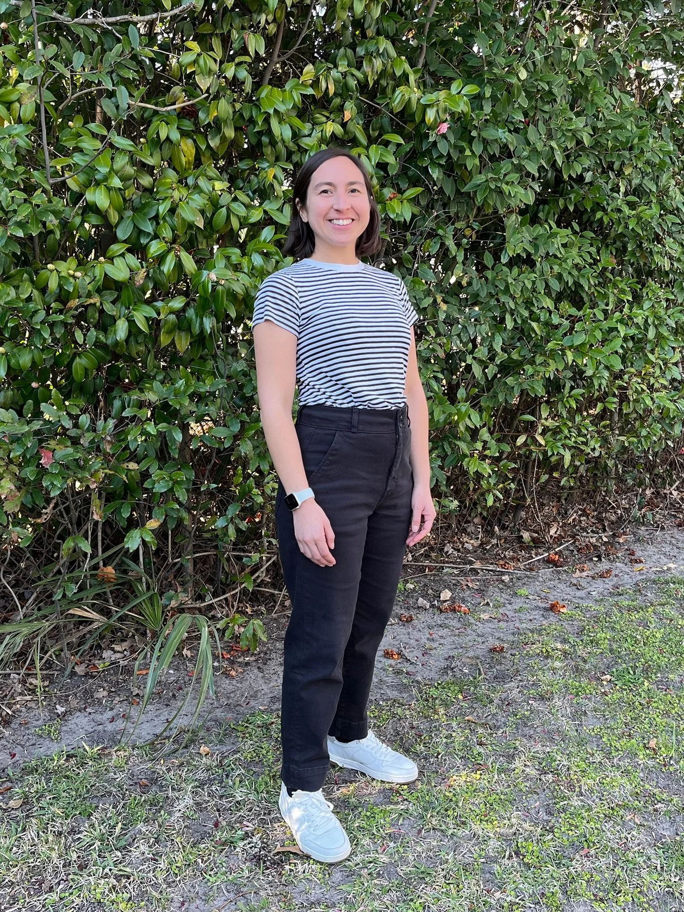 A woman smiling outdoors standing in front of green leafy bushes, wearing a striped t-shirt, black pants, white sneakers, and a smartwatch on her left wrist.