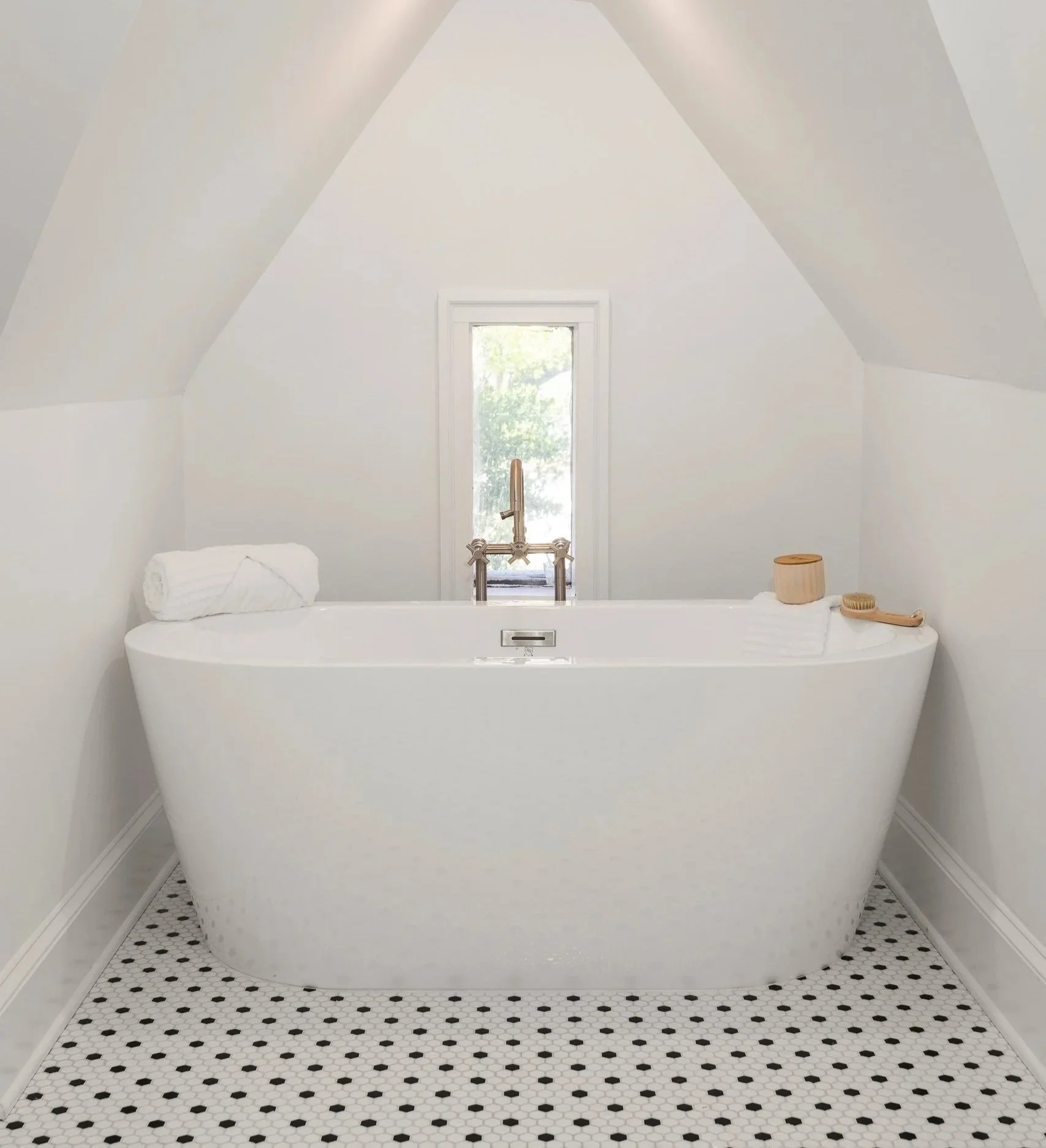 White freestanding bathtub with a rolled towel on the edge, set in a small bathroom with white walls, a small window behind the tub, and black and white hexagonal floor tiles.