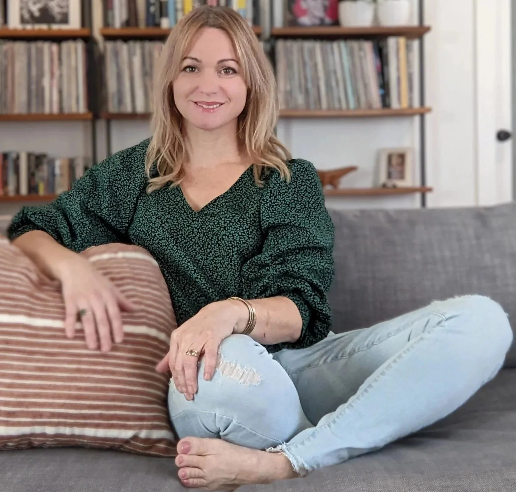 A woman sitting on a gray sofa with her legs crossed, smiling at the camera, in a room with bookshelves filled with books in the background.