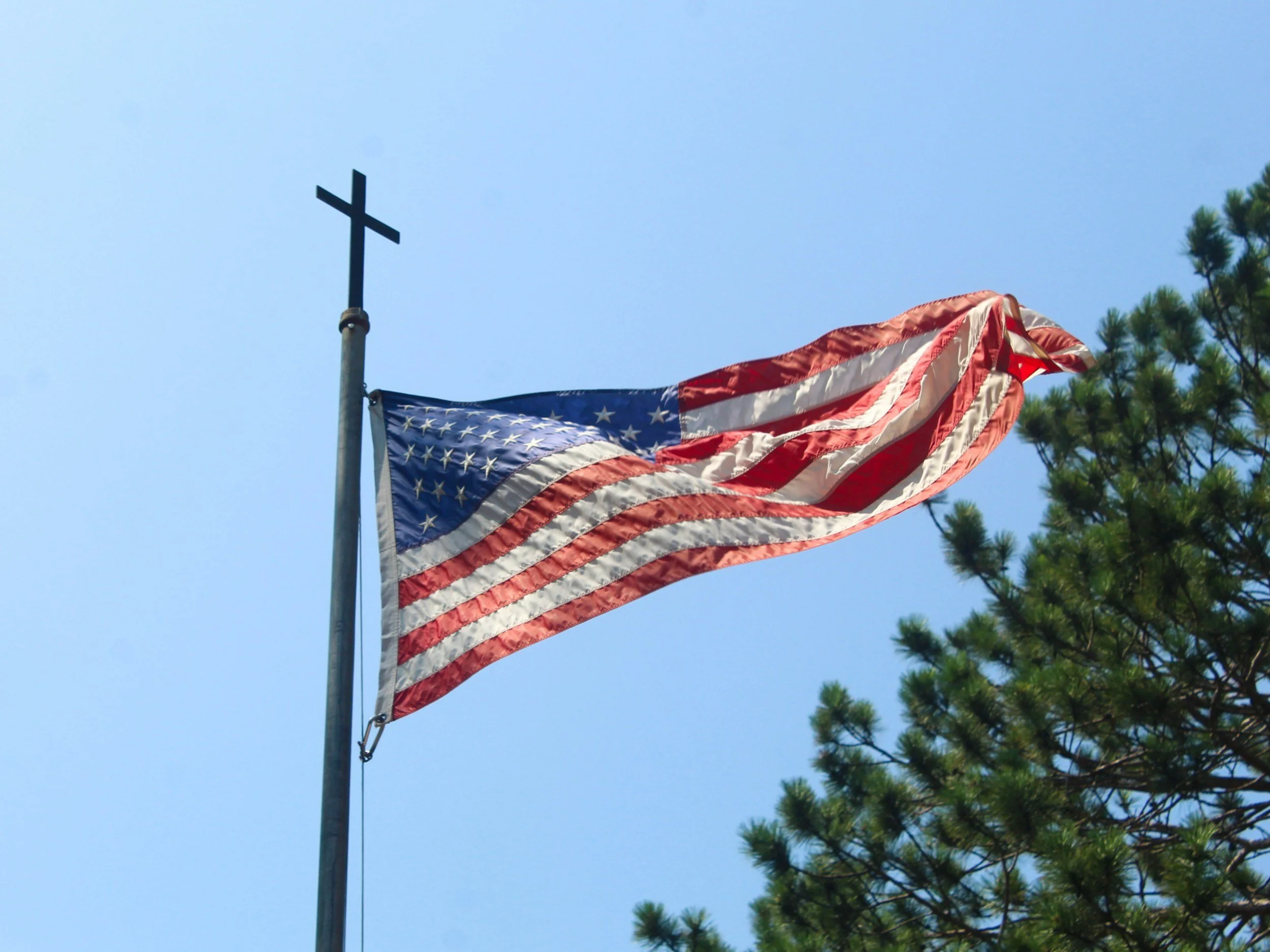 American flag flying on a pole with a blue sky and green trees in the background.