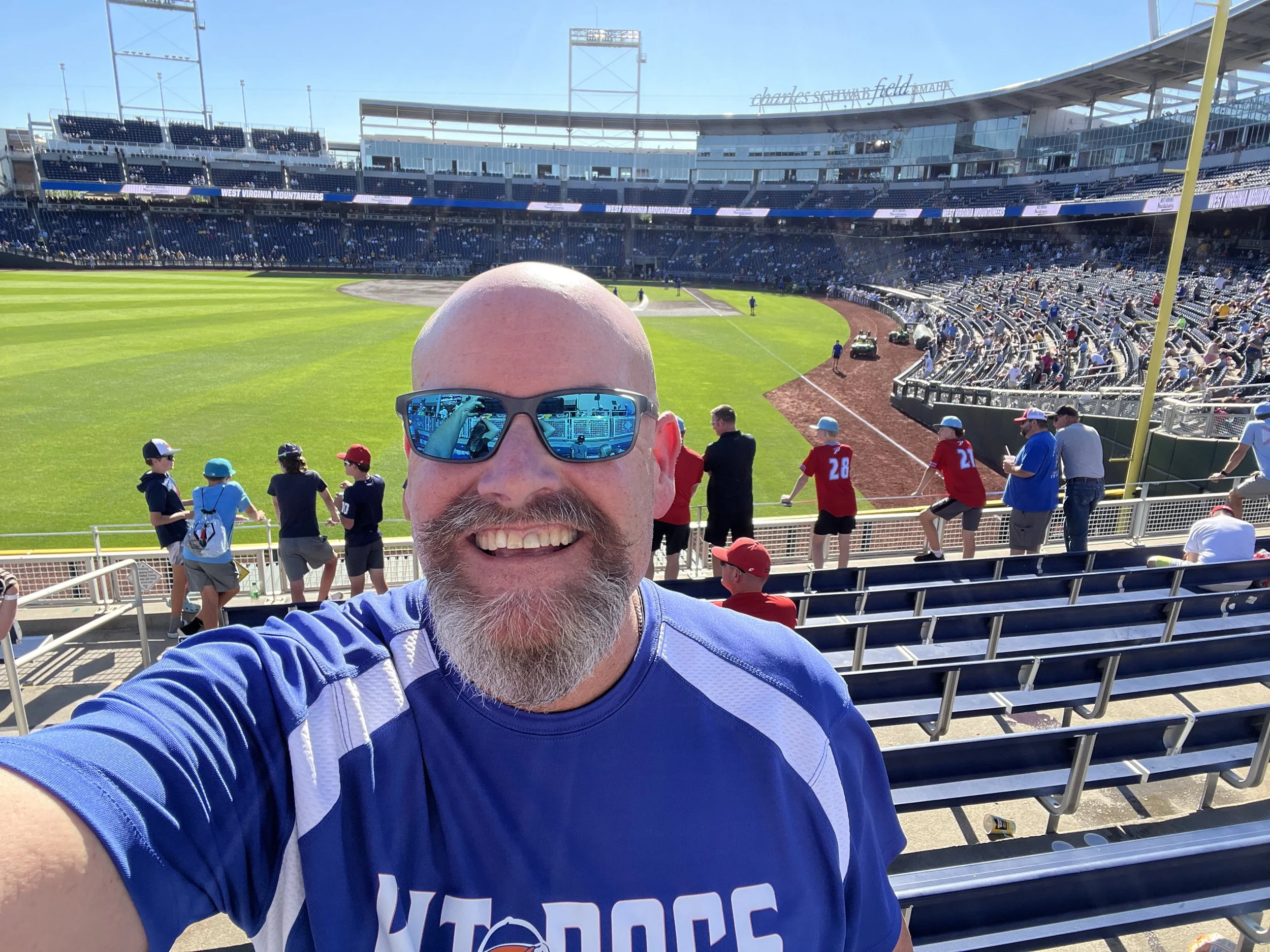 The HR-VET with his bald head, big smile, goatee, and sunglasses is taking a selfie. The background shows a bright sunny day at the Charles Schwab Baseball Field filled with spectators and players on the field.