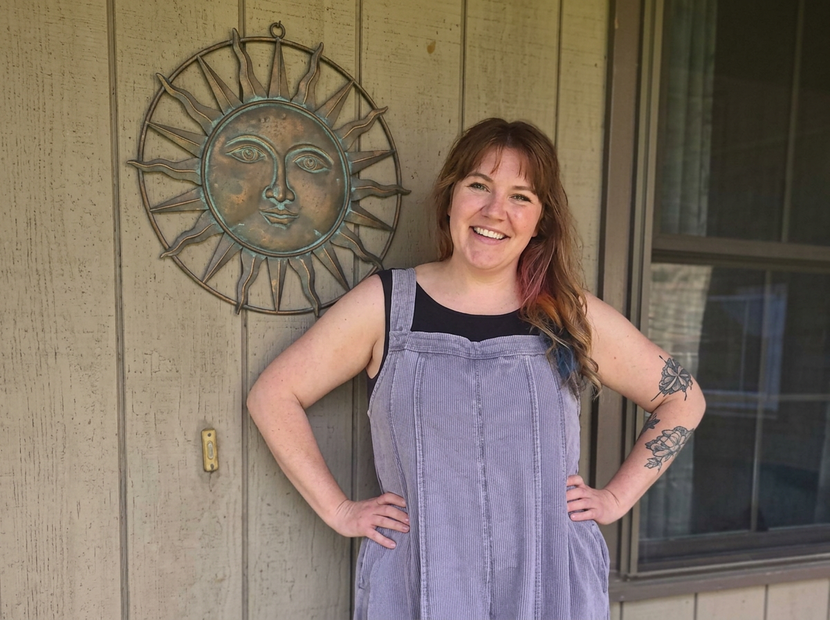 A woman with long hair and floral tattoos on her arms smiling with hands on hips, standing outside next to a house wall with a sun-shaped decorative metal piece with a face behind her.