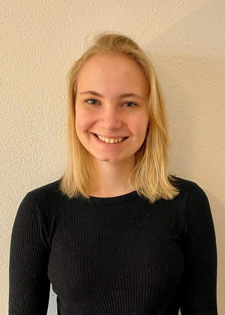 Ein junges Mädchen mit blondem, schulterlangem Haar, das fröhlich lächelnd vor einer hellen Wand steht.