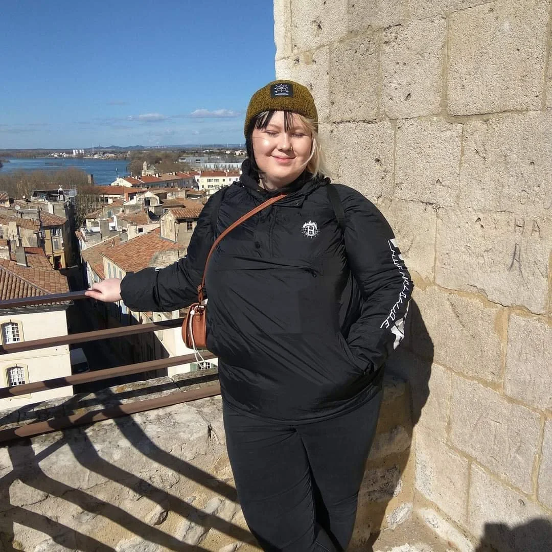 Junge Frau in dunkler Jacke und grüner Mütze steht an einer Burgmauer mit Blick auf eine Altstadt und einen Fluss unter blauem Himmel.