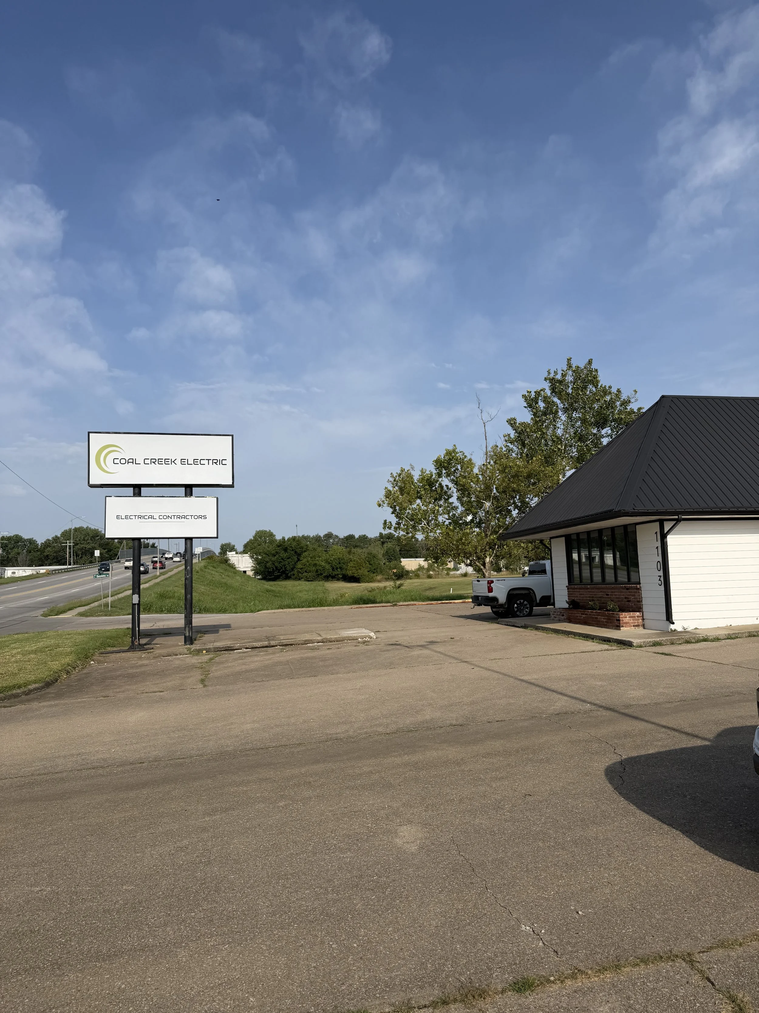 A clear day with a blue sky and scattered clouds. In the foreground, there is a parking lot with a white pickup truck parked near a small building with a dark roof. To the left, there are two signs: one indicating the business name "Coal Creek Electric" and the other indicating "Electrical Contractors." There are some trees and a highway in the background.