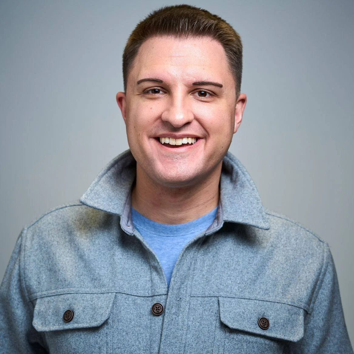 A smiling young man with short brown hair, wearing a light gray jacket with a collar over a blue shirt against a plain gray background.