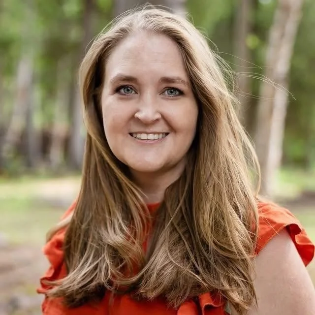 A smiling woman with long, wavy blonde hair, wearing an orange top, standing outdoors with trees in the background.