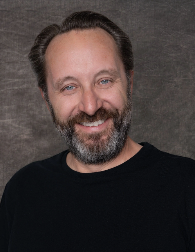 A middle-aged man with a beard and light blue eyes smiling against a gray textured background.