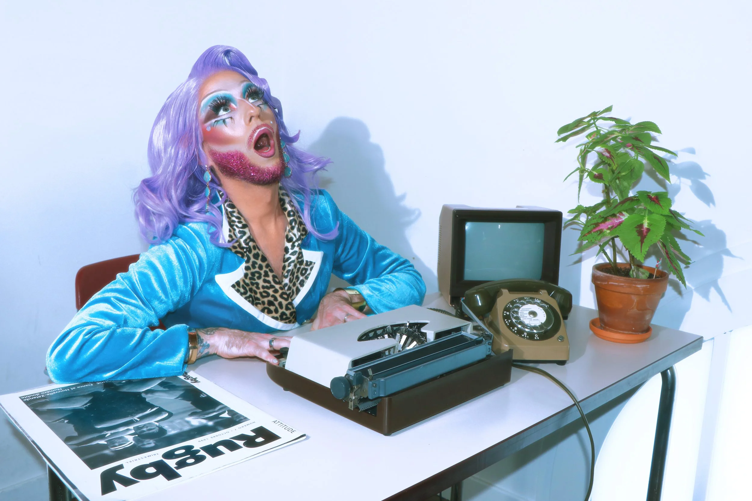 Une personne avec maquillage extravagant, cheveux violet, portant une veste bleue, est assise à un bureau avec une vieille télé vintage, un téléphone rotatif, une plante en pot, et des magazines.