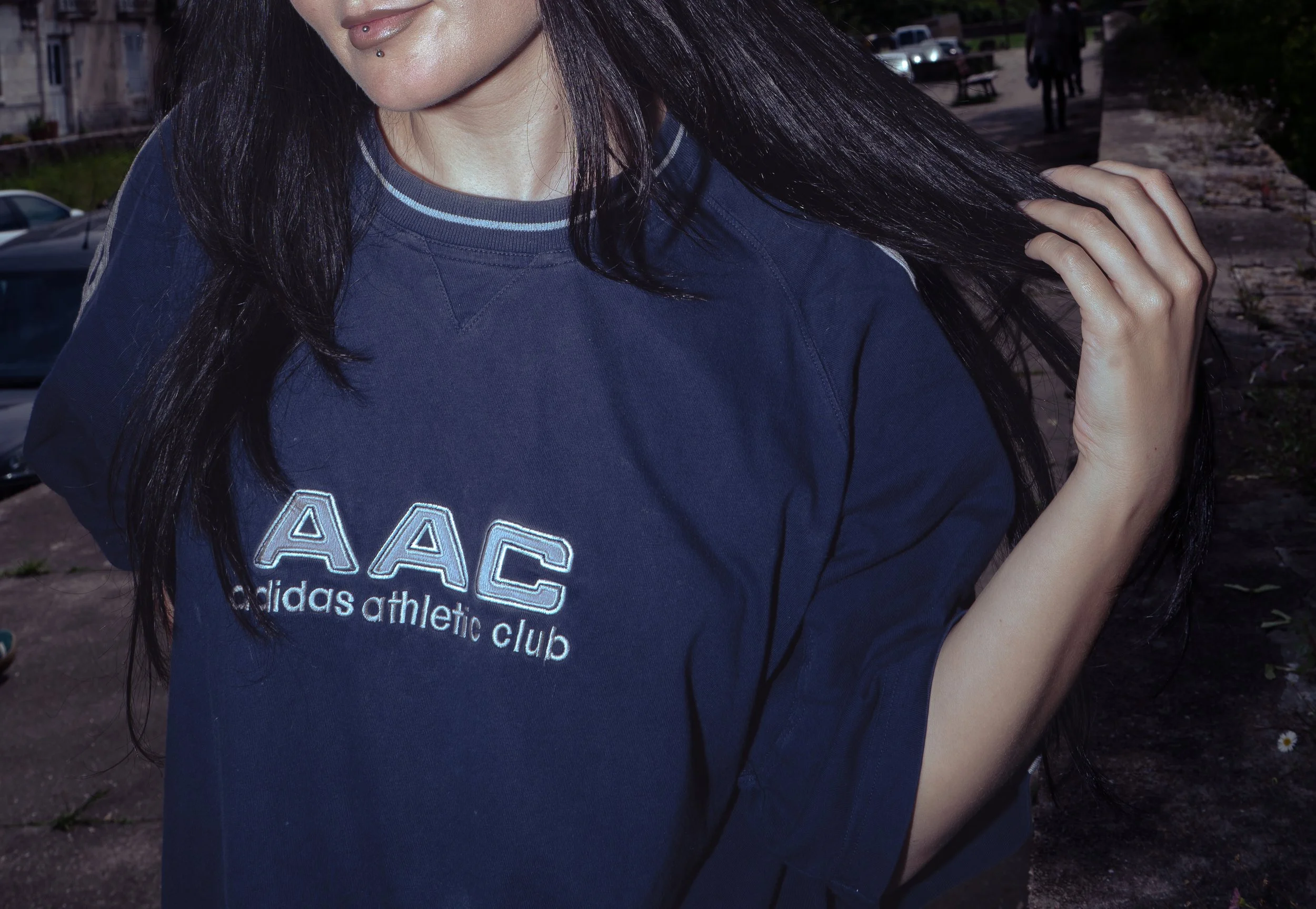 Une femme avec des cheveux longs noirs portant un sweat-shirt bleu marine de l'Adidas Athletic Club, se tenant à l'extérieur en soirée. Elle a des piercings sur la lèvre inférieure. Le fond montre des voitures, un banc et deux personnes marchant, avec une ambiance nocturne.