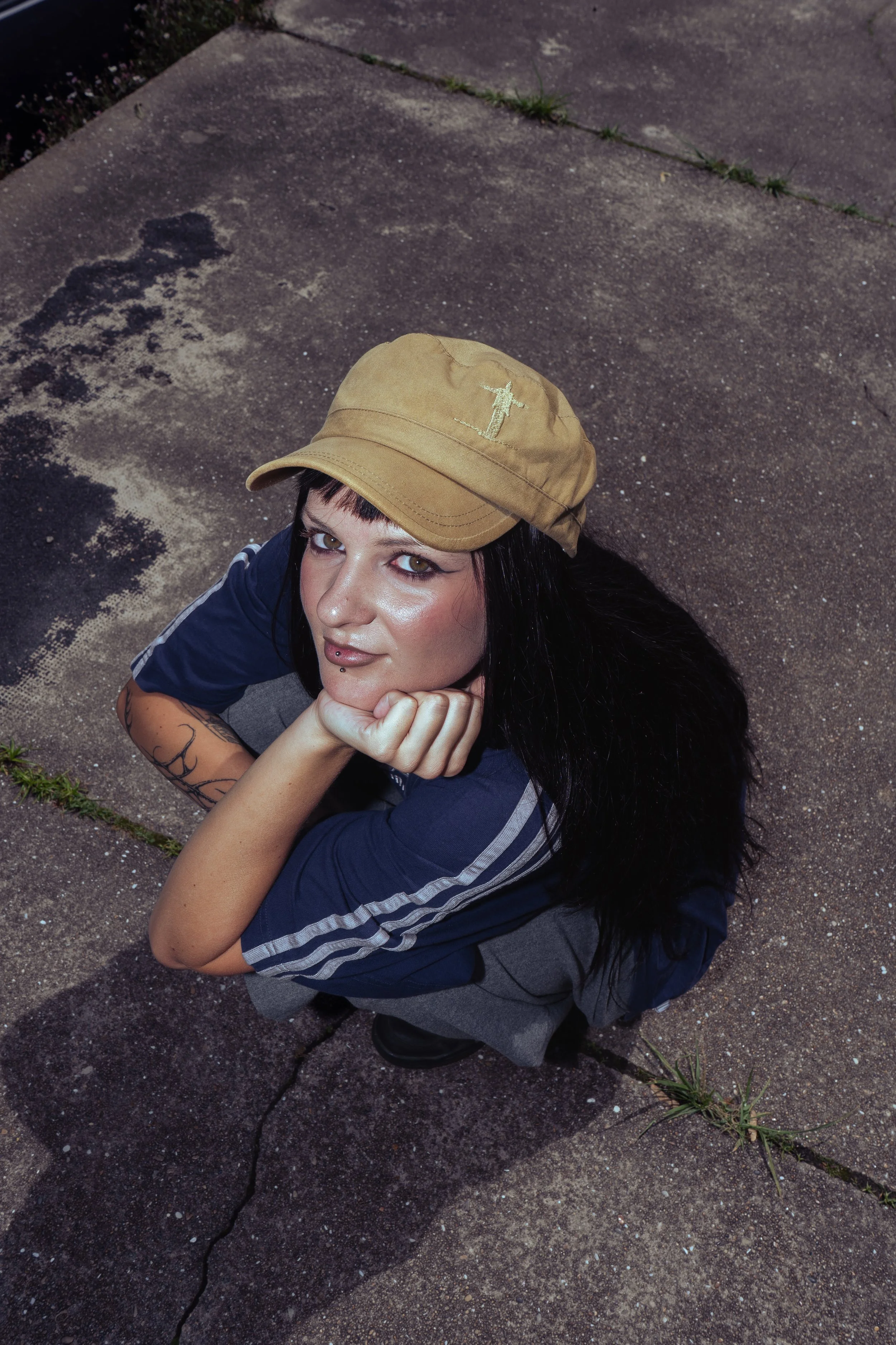 Jeune femme assise sur un trottoir en béton, portant une casquette beige, une chemise bleue avec des rayures blanches sur les manches, regarde vers le haut avec un sourire, des tatouages sur le bras gauche, cheveux longs et noirs, décor extérieur avec de l'herbe poussant à travers une fissure sur le trottoir.