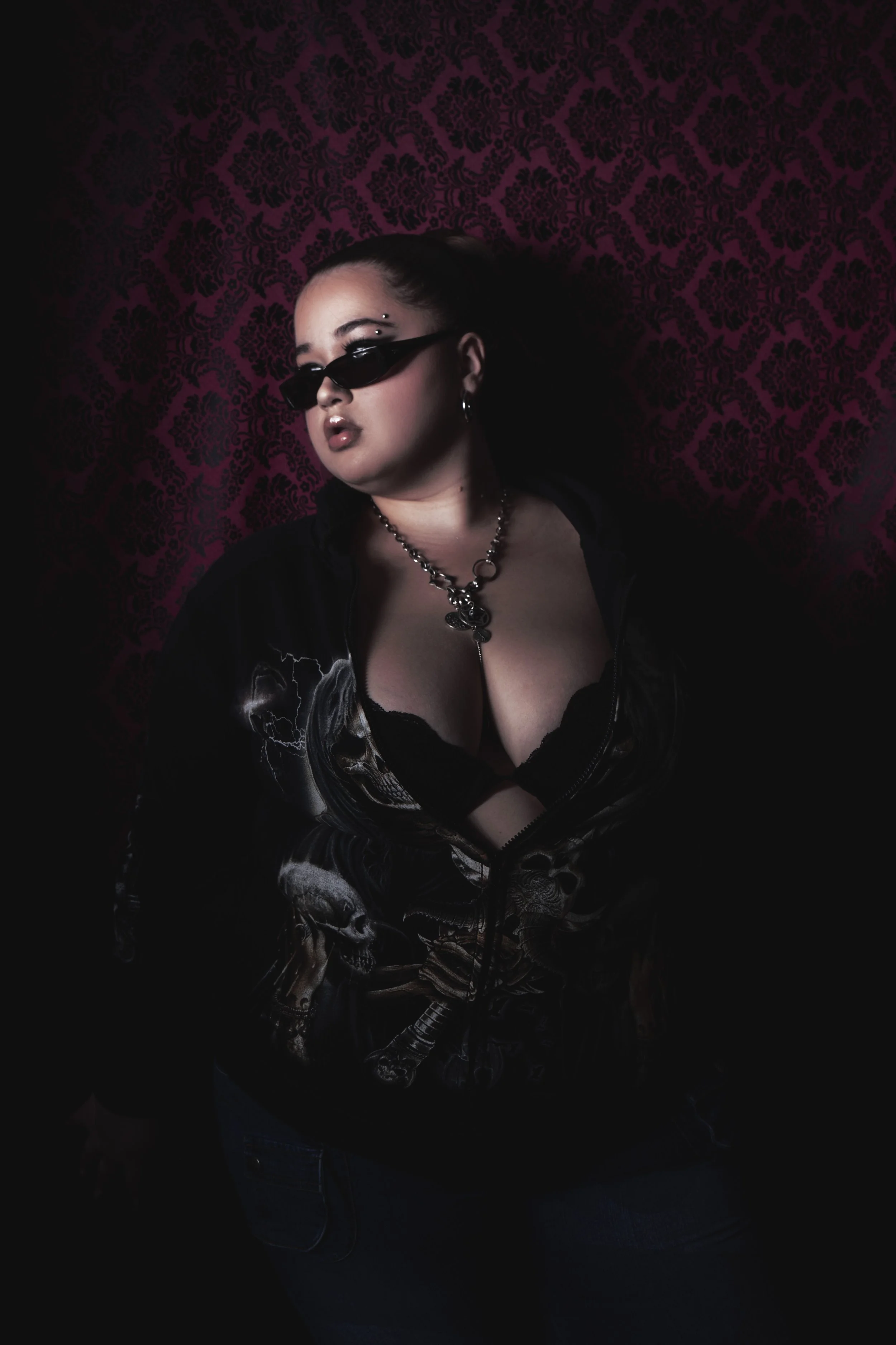 Une femme portant des lunettes de soleil noires, un collier en chaîne, une veste en cuir avec des motifs skulls, pose devant un fond violet avec un motif noir, dans un style gothique.