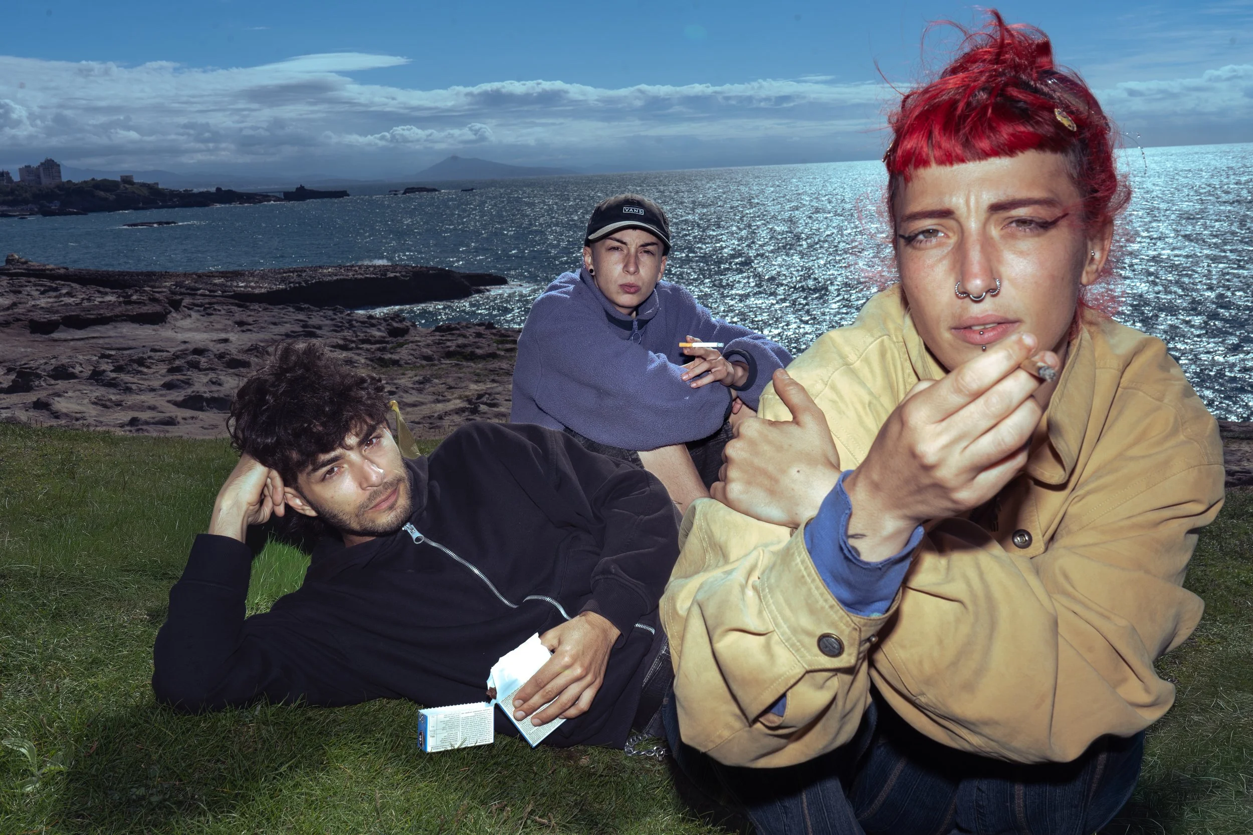 Trois jeunes gens au bord de la mer, un homme allongé portant une veste noire, une femme avec des cheveux roux tenant une cigarette, et un jeune homme en sweat bleu portant une casquette, avec un paysage marin en arrière-plan.