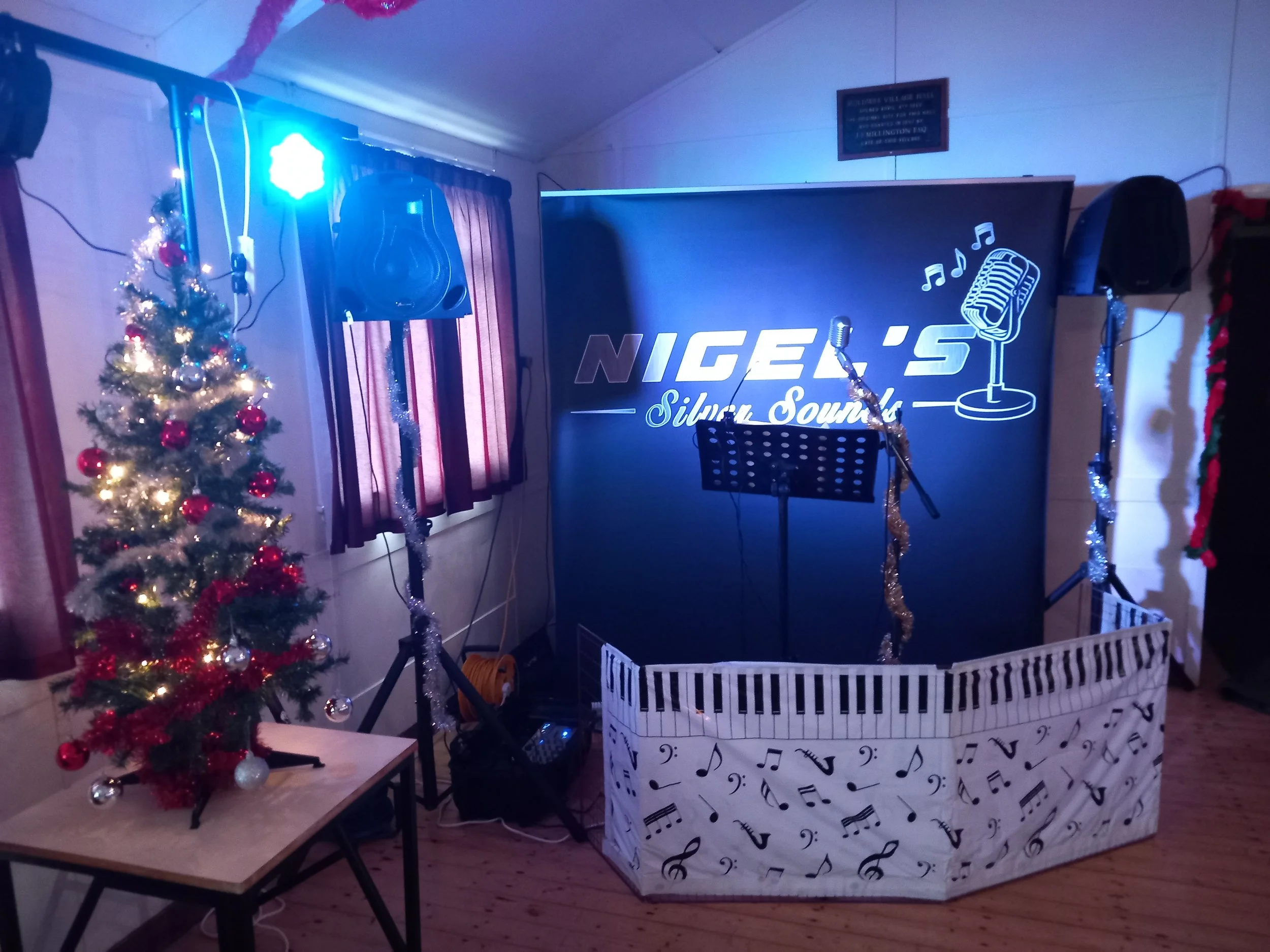 A festive indoor scene with a decorated Christmas tree adorned with red and silver ornaments and white lights, a small black music stand with a sheet of music, a stage with a black backdrop featuring the words "NIGER'S Silver Sounds" with musical not