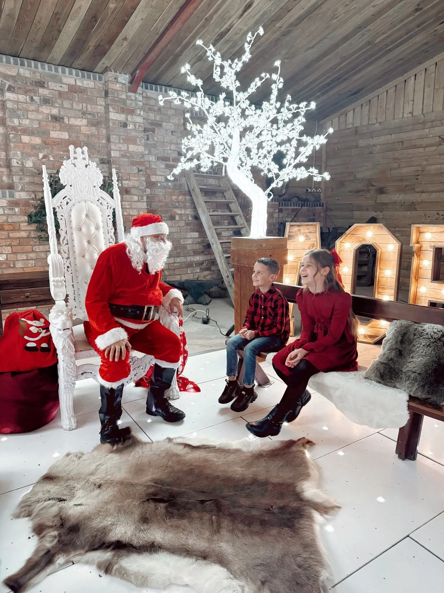 Santa Claus in a red suit and hat sitting on a white ornate throne in a rustic indoor setting, talking to two children who are sitting on a bench. Two large animal pelts are on the floor in front of them, and a lit decorative tree is visible in the b