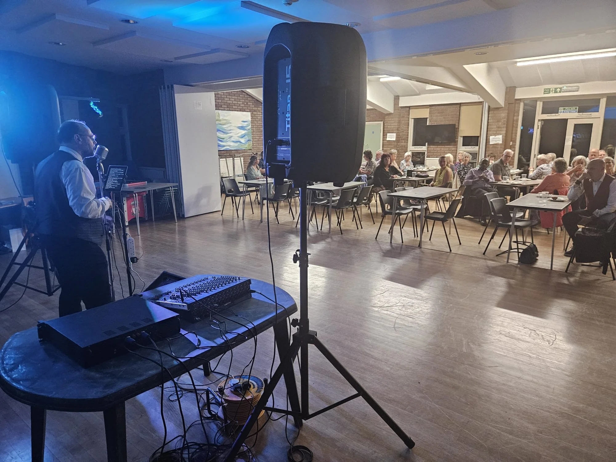 A performer with short hair, glasses, a white shirt, and a dark vest stands on stage with a microphone and music stand, while a large speaker and DJ equipment with cables are in the foreground. The audience, consisting of mostly elderly people, is se