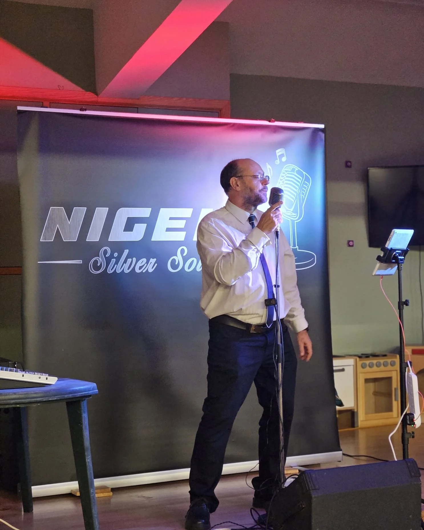 Male performer in a white shirt, black tie, and black pants holding a microphone on stage with a backdrop that says 'NIGER Silver S' and musical graphics.