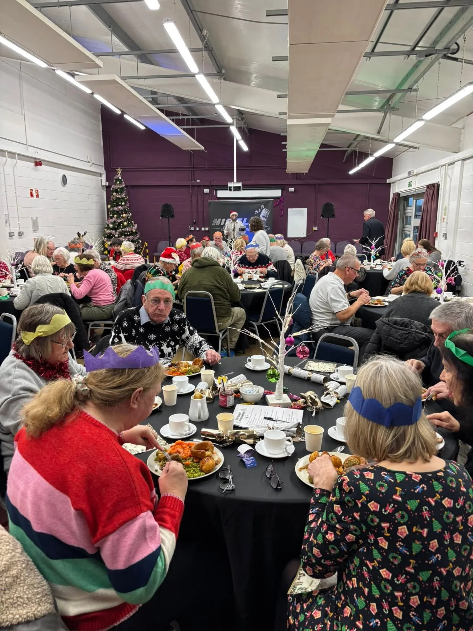 People gathered around tables during a festive holiday celebration, with Christmas hats and reindeer antlers, decorated with Christmas ornaments and a Christmas tree, in a community hall with purple walls and stage in the background.