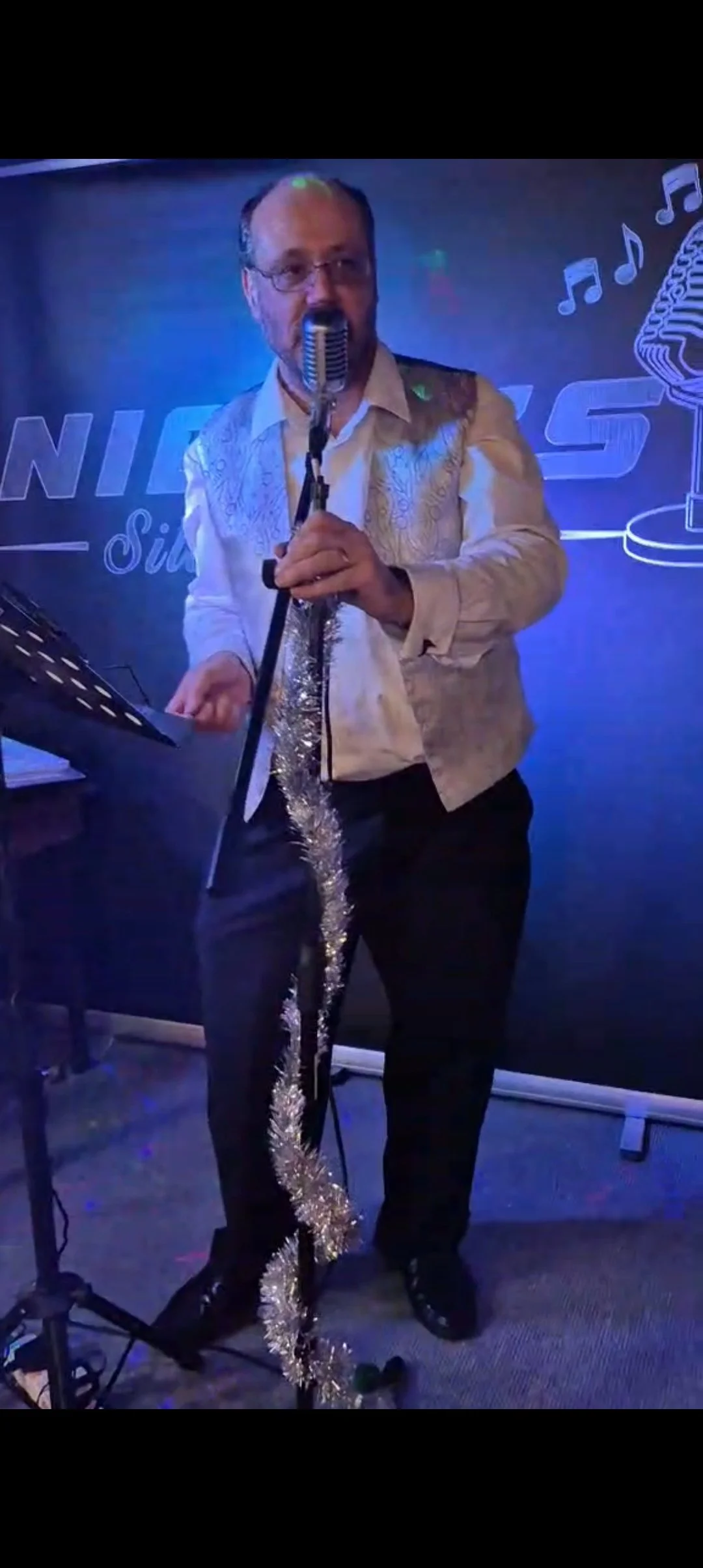 A man in a light-colored, embroidered shirt holding a microphone decorated with silver tinsel, standing in front of a dark blue backdrop with musical notes and a microphone graphic, at a singing performance or karaoke event.