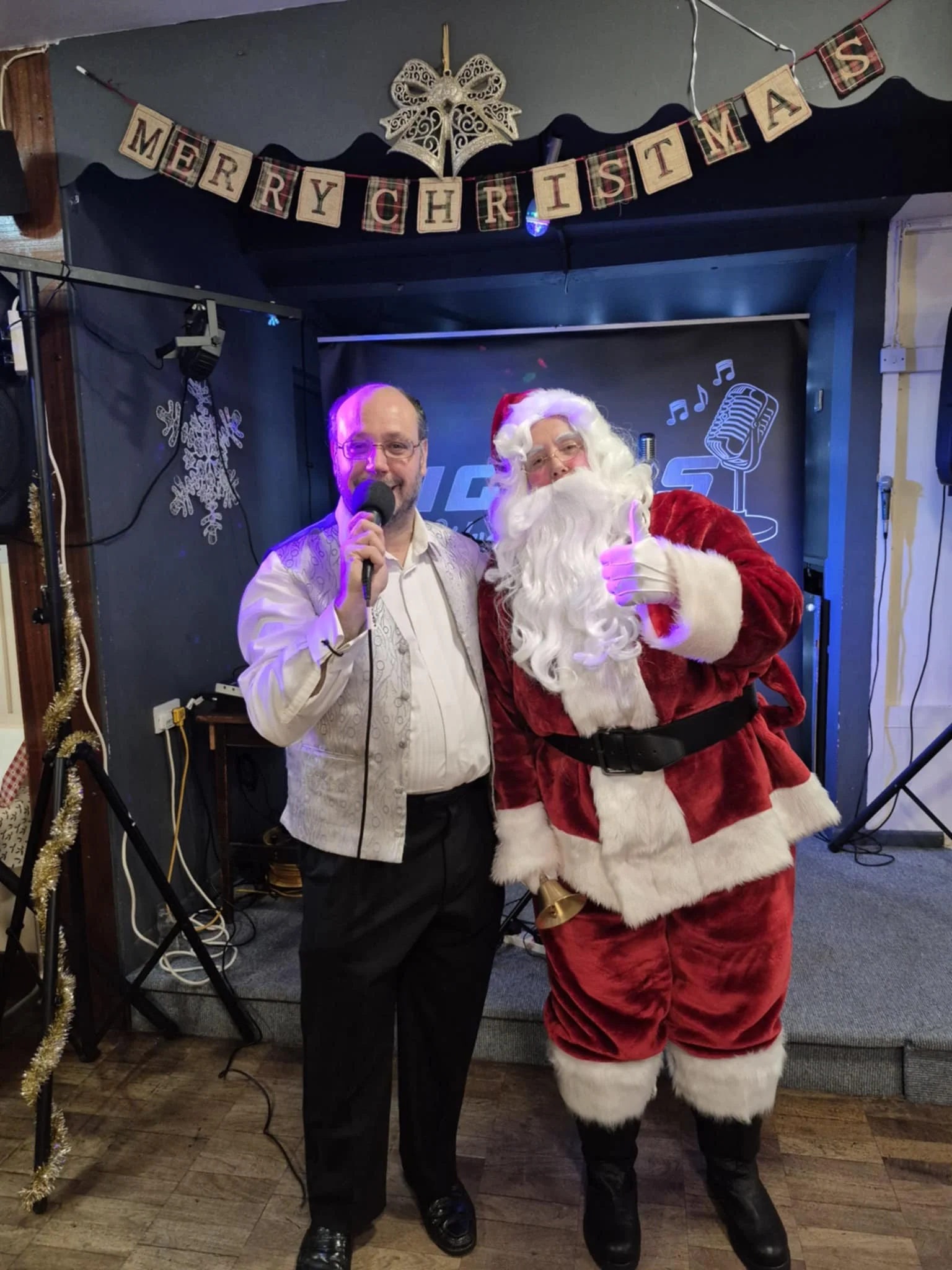 Man and person dressed as Santa Claus standing together at a Christmas celebration with a 'Merry Christmas' banner above them.