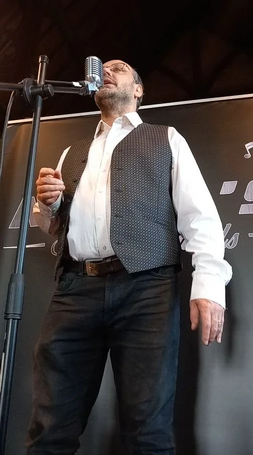 A man singing or speaking into a microphone on stage, wearing glasses, a white shirt, a black vest, and dark pants, with a dark background and stage lighting.