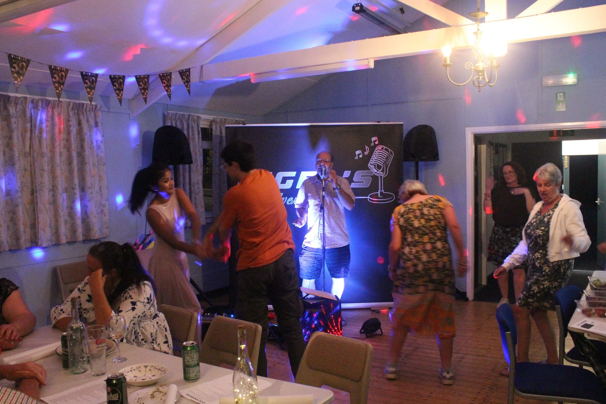 People dancing and socializing at an indoor celebration or party with a live performer singing on stage, decorated with a banner that says '50', and colorful lighting.