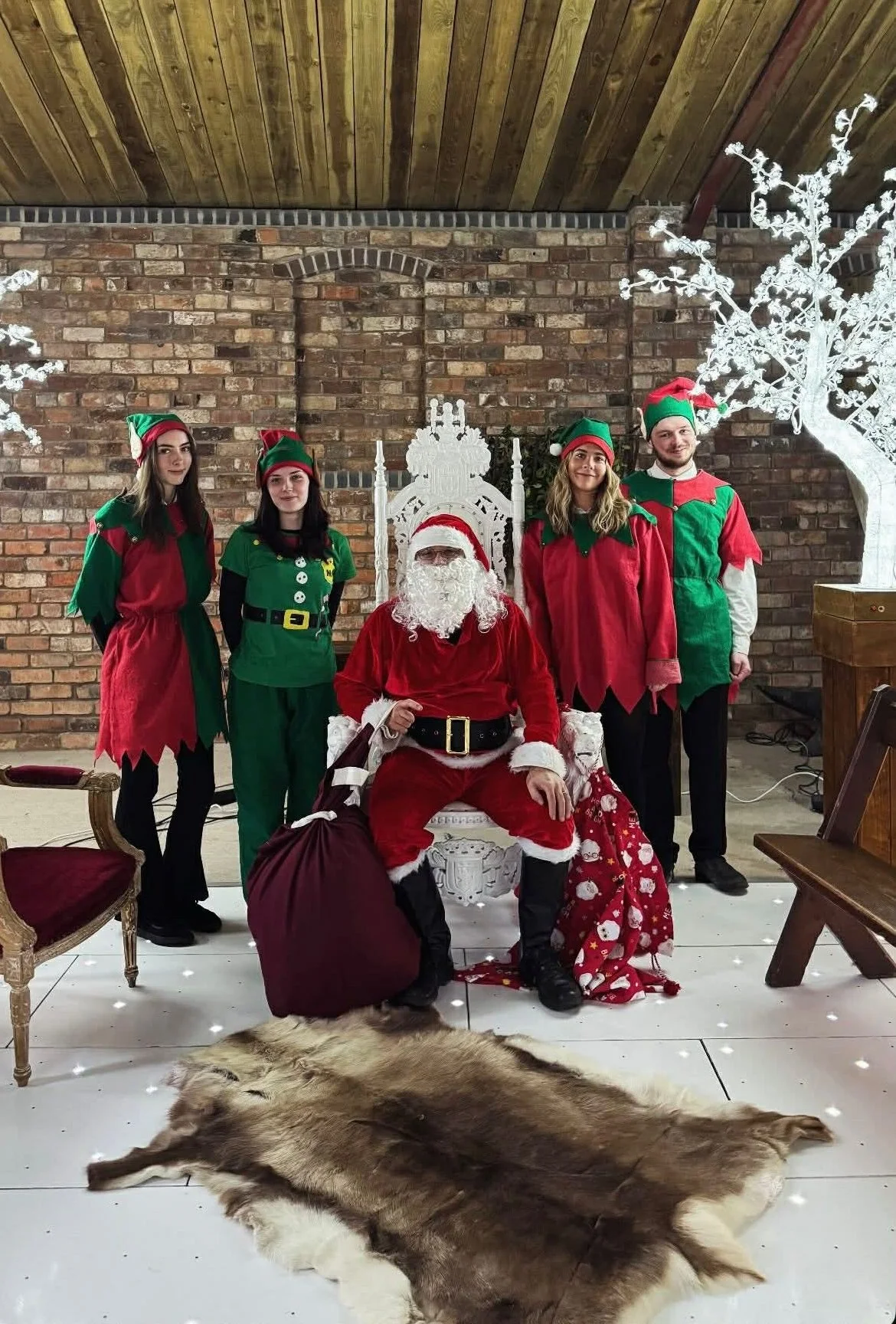 Group photo with Santa Claus and four people dressed as elves or Christmas characters, seated on a decorative white chair with a red pillow, in a festive setting with a brick wall background, white Christmas trees, and a fur rug on the floor.
