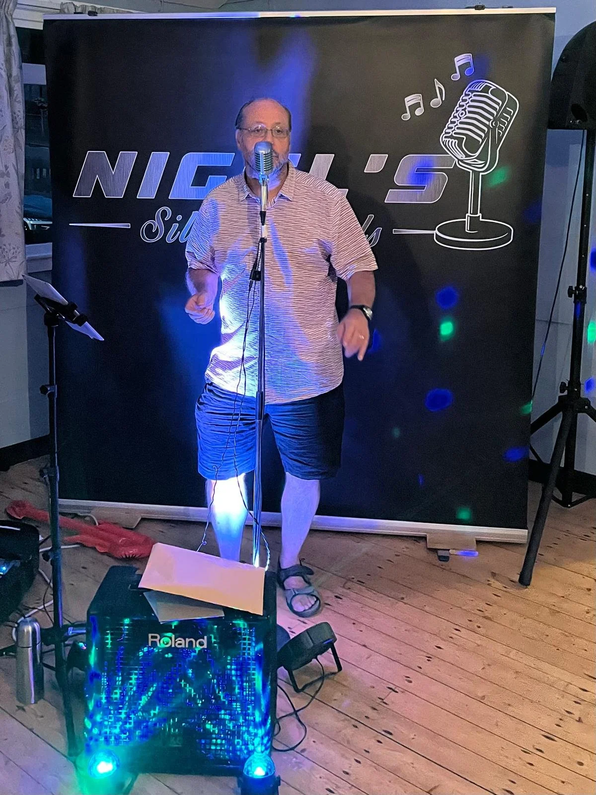 A man singing into a vintage microphone on a small stage with a black backdrop that has a logo reading 'NICK 'S' and a drawing of a microphone with musical notes around it. The man is wearing a striped short-sleeved shirt, shorts, sandals, and a watc