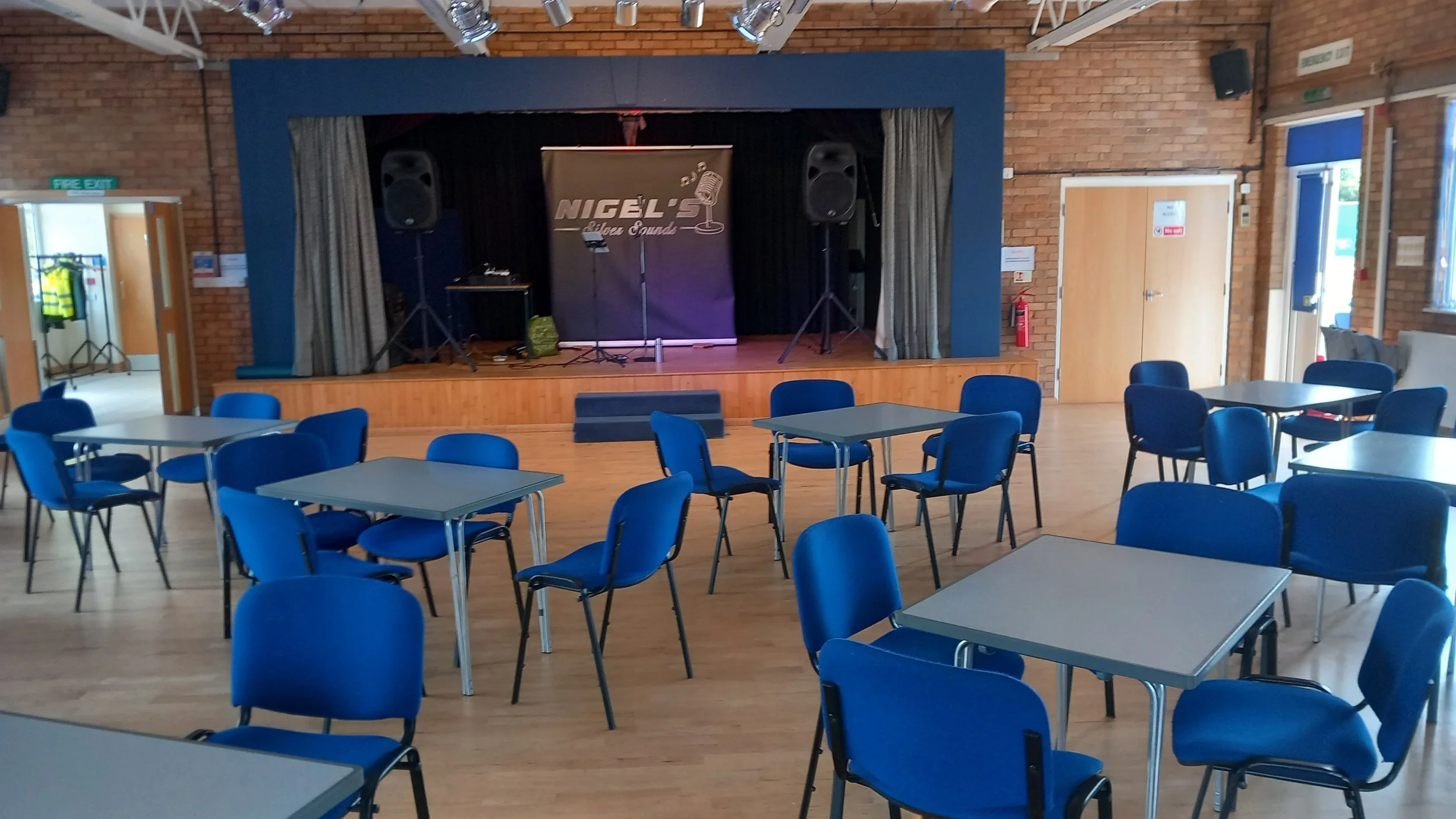 An empty stage with a microphone, speakers, and a backdrop with the text 'Nicel's' and a graphic of a microphone. In front of the stage, there are multiple tables and chairs arranged in a room with brick walls and windows.
