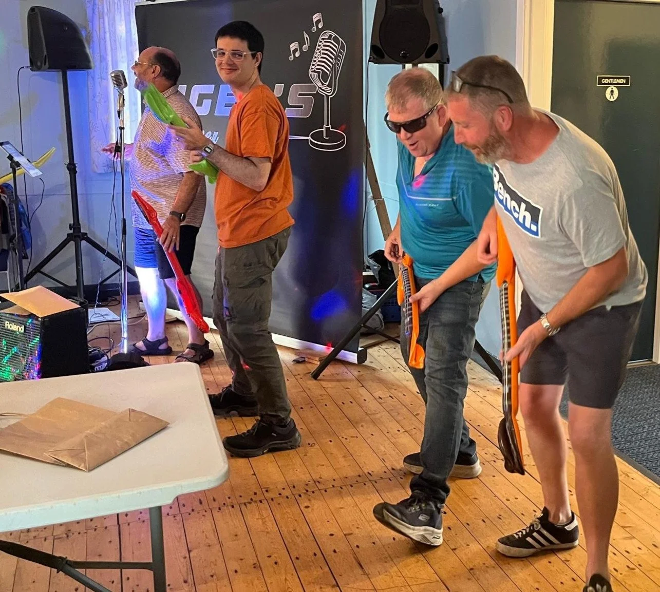 Four men standing in a row holding guitars, on a wooden floor, performing or practicing music. They are near a microphone, speakers, and a banner with a microphone graphic. One man wears sunglasses, and others wear casual t-shirts and shorts.