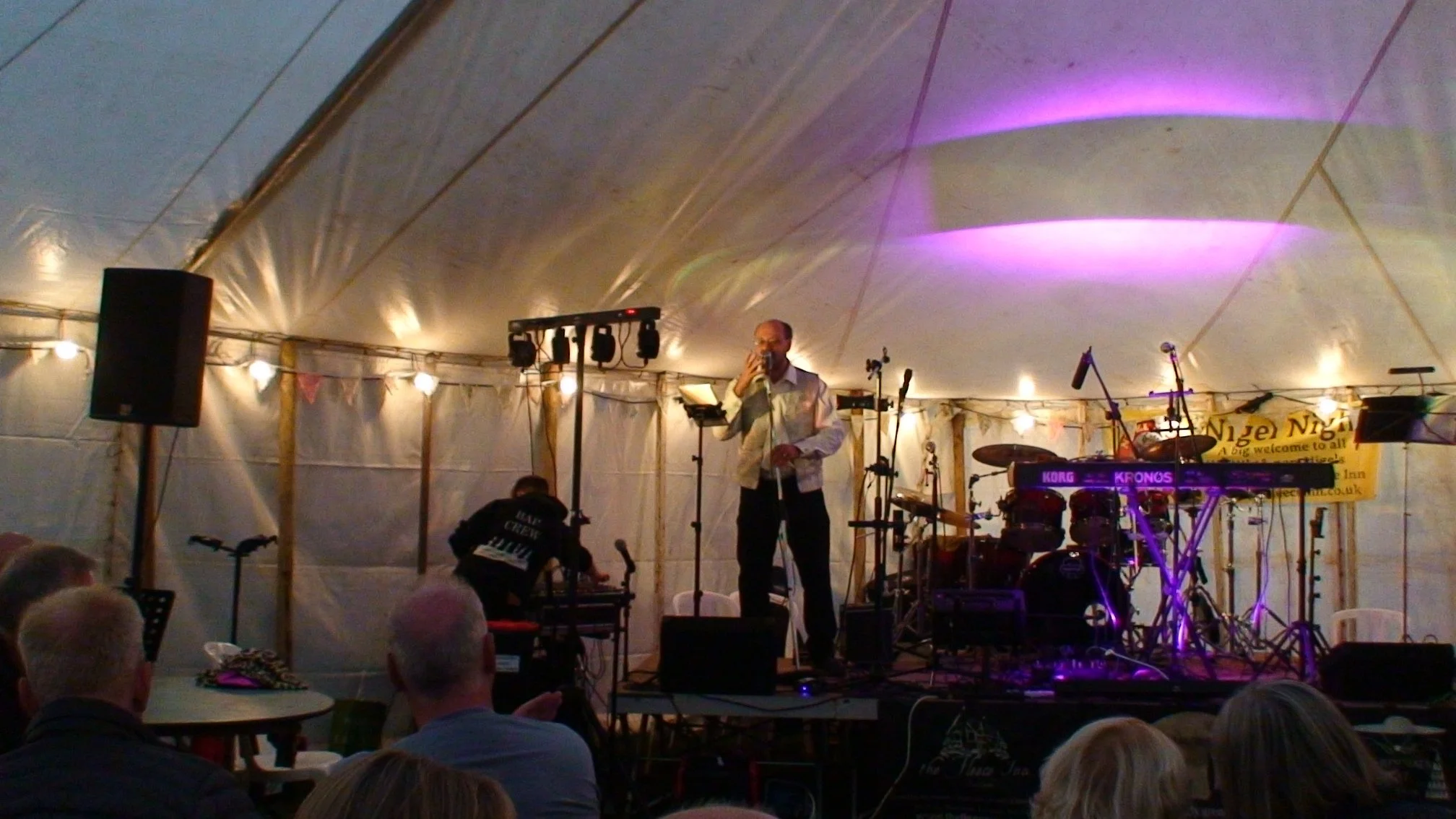Man with a microphone on stage in a large white tent, musical instruments including drums and microphones around him, audience seated in front with heads visible.