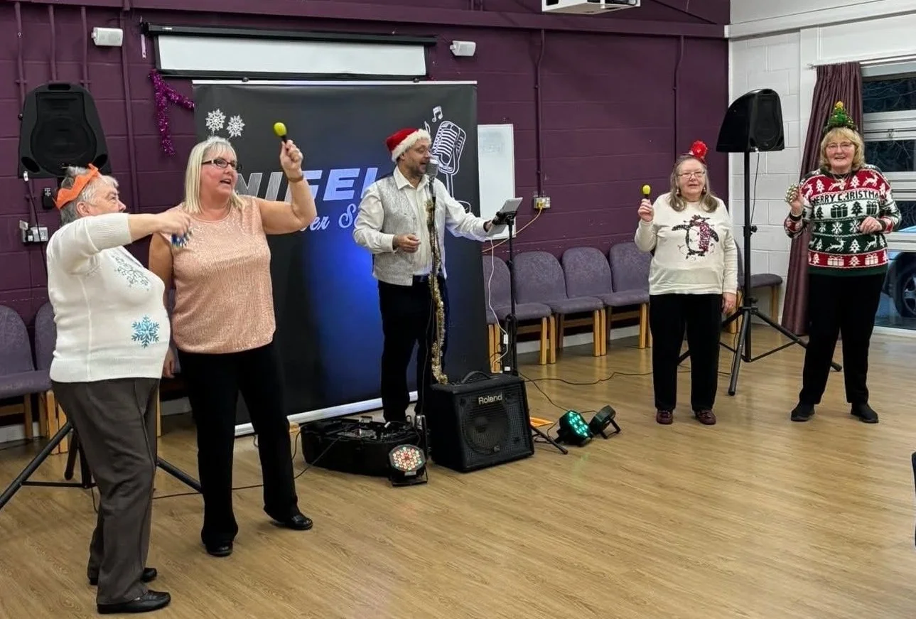 Group of five women and one man singing or performing at a Christmas party, wearing festive sweaters and accessories in a decorated room.