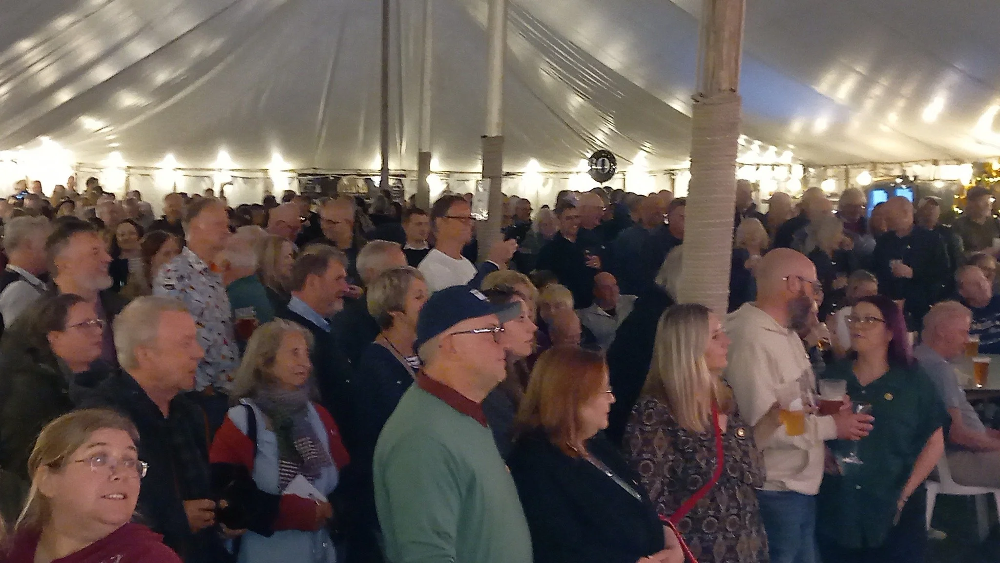 Crowd of people attending an indoor event under a large white tent with string lights and white brick columns.
