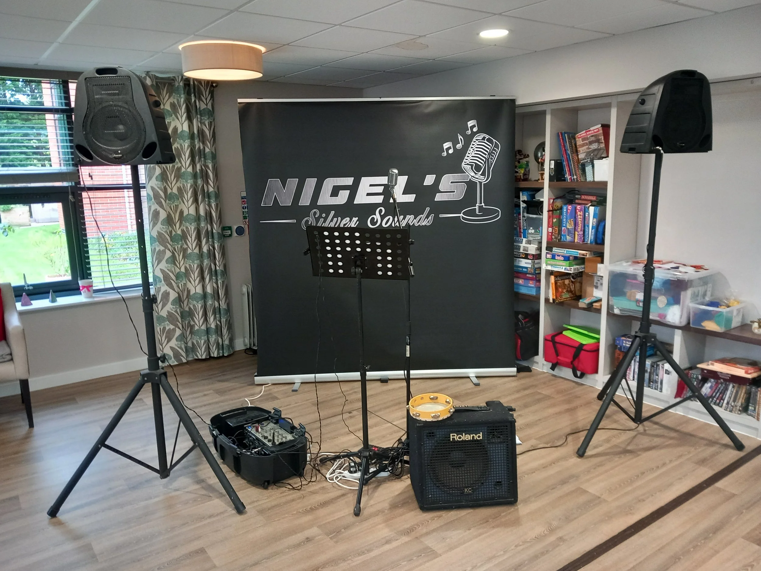 A room set up for music or recording, with two large black speakers on stands, a microphone on a stand, a music stand, and various equipment and cables on the floor. There is a backdrop with "NIGEL'S Silver Sounds" and musical notes and a microphone 
