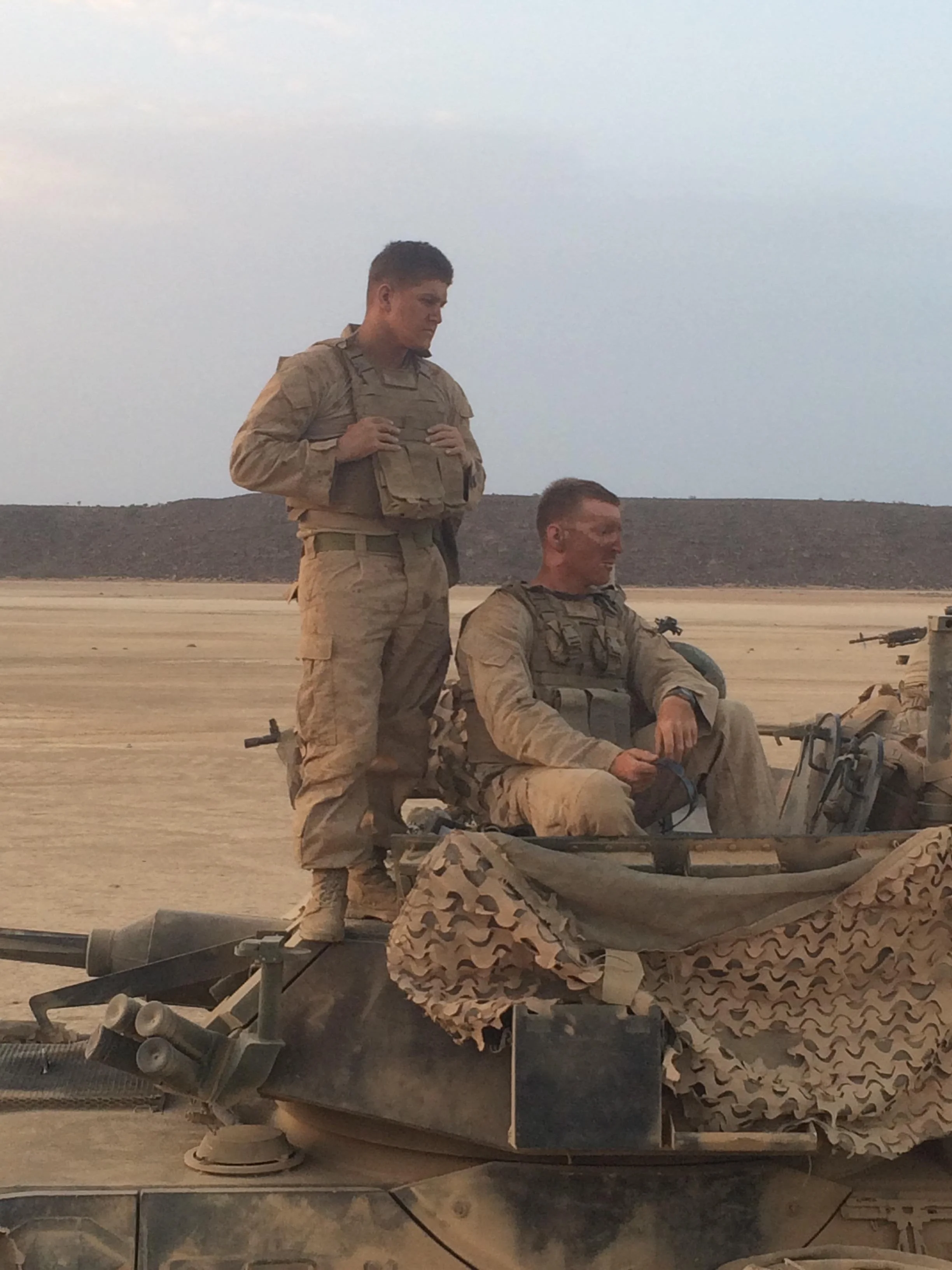 Two Marines in desert camouflage uniforms, one standing and one sitting on a military vehicle, in a barren desert landscape with a cloudy sky.