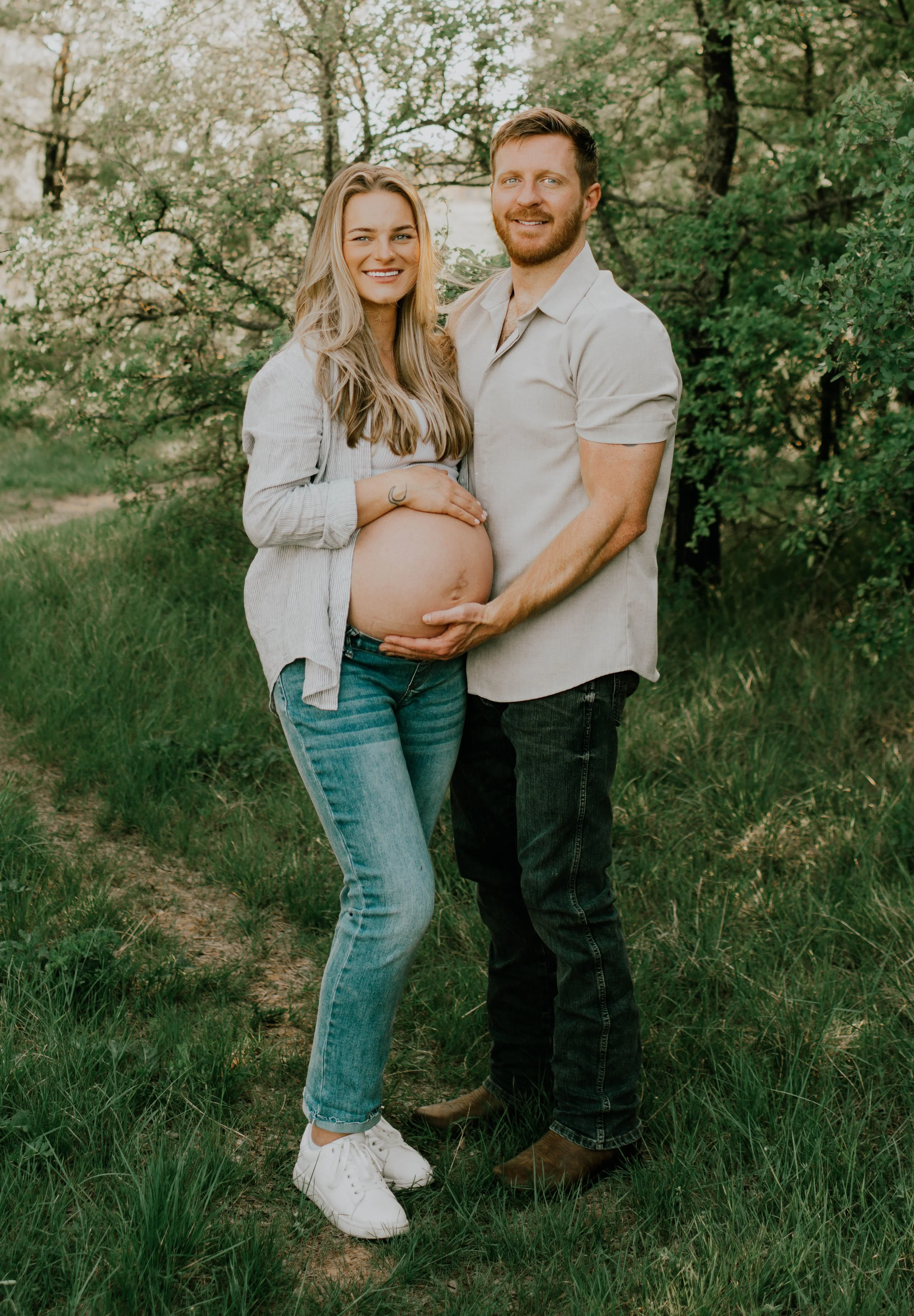 A pregnant woman and a man are standing outdoors in a grassy area with trees in the background, smiling and holding her belly.