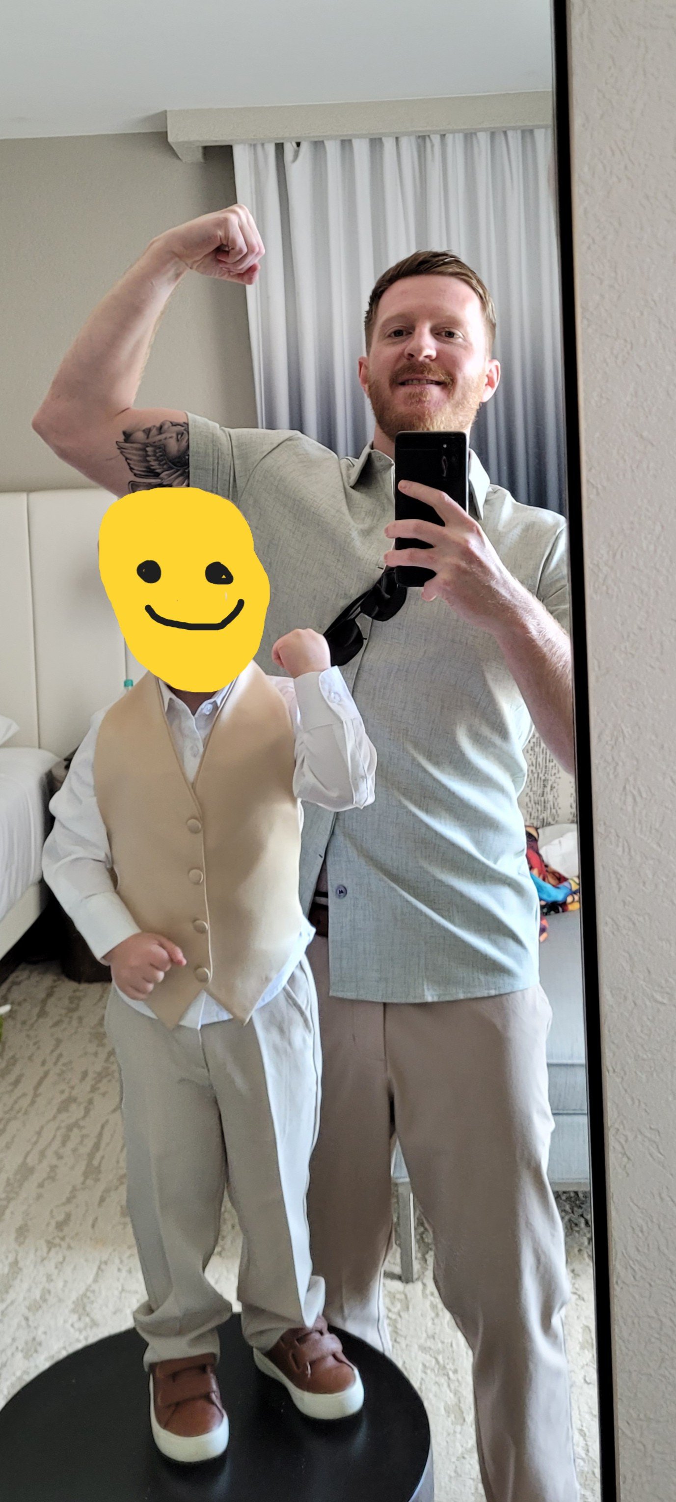 A man taking a selfie in a hotel room mirror, flexing his arm, and a child with a yellow smiley face sticker covering their face, dressed in formal attire, standing on a black circular pedestal.
