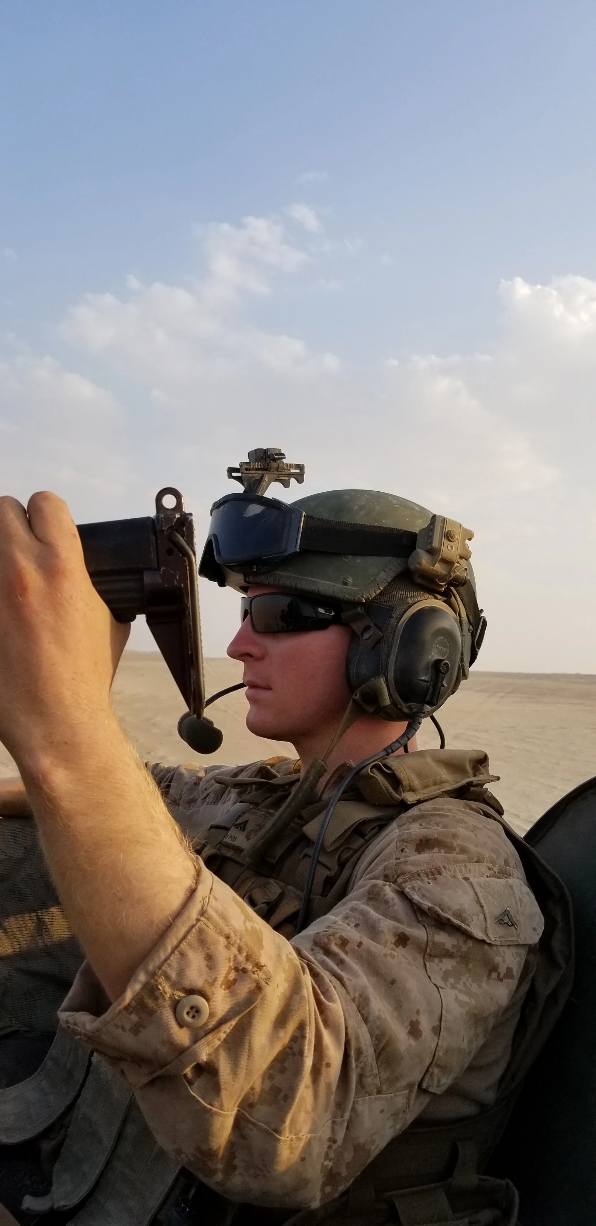 A Marine in camouflage uniform, wearing tactical gear and a helmet with  goggles, is aiming a rifle in a desert landscape under a partly cloudy sky.