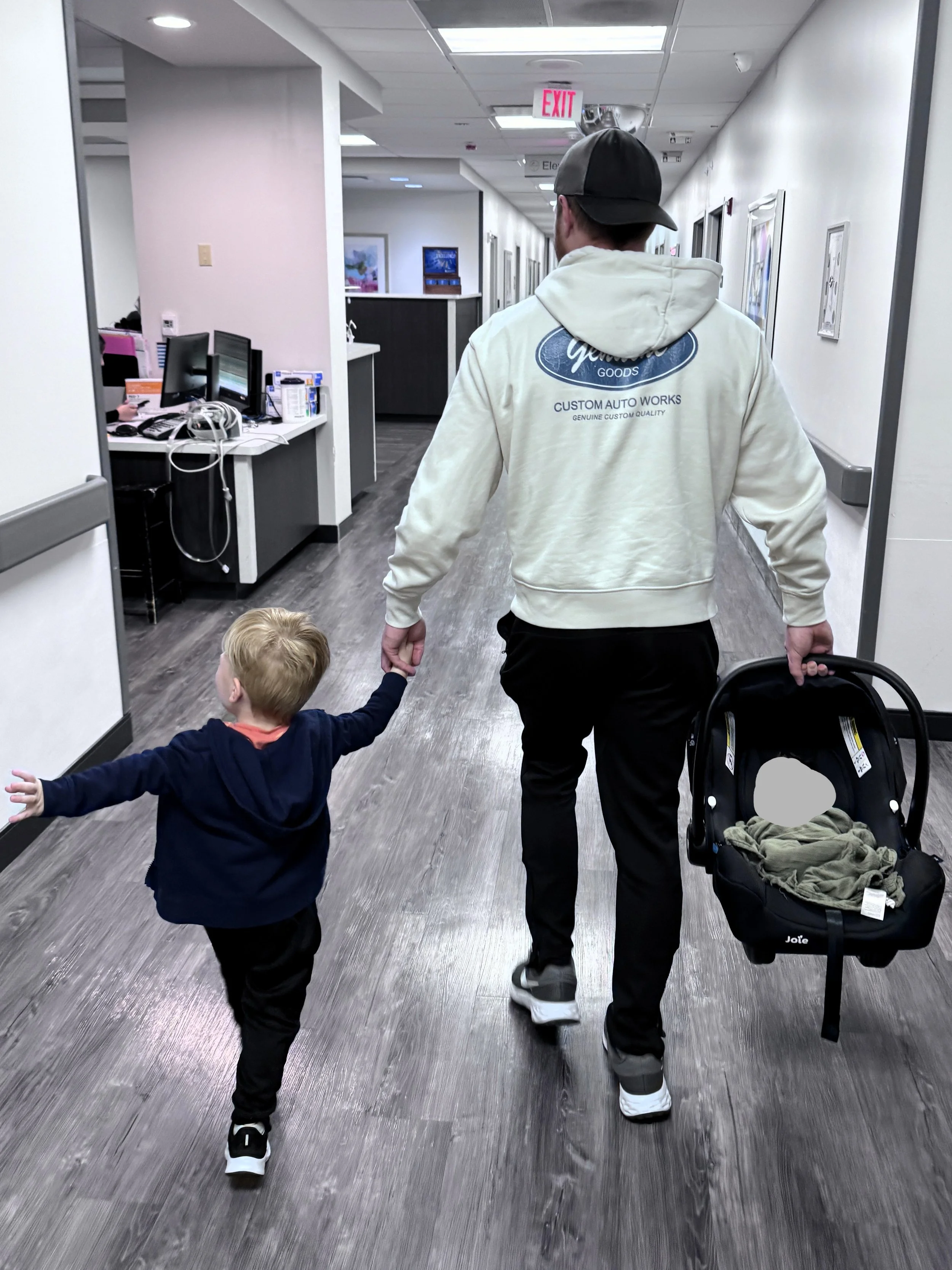 A man and two children walking down a hospital corridor. The man is holding a stroller and a child's hand. The corridor has gray flooring, white walls with framed pictures, and a row of workstations with monitors on the left. The man is wearing a baseball cap, a light-colored hoodie, and black pants, while the children are dressed in dark clothing.