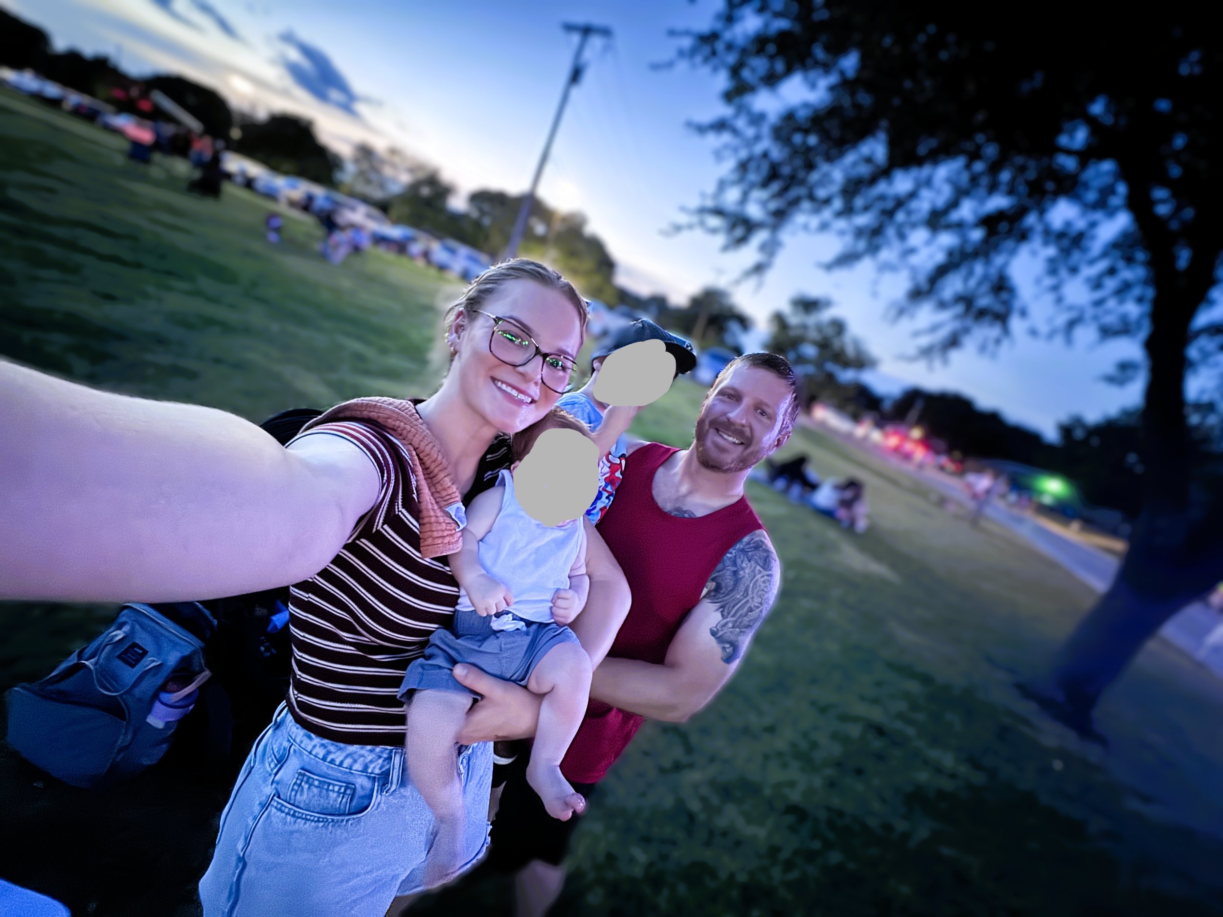 Smiling woman wearing glasses and a striped shirt taking a selfie with two men and children at a park during evening, with a blurred background of trees and parked cars.
