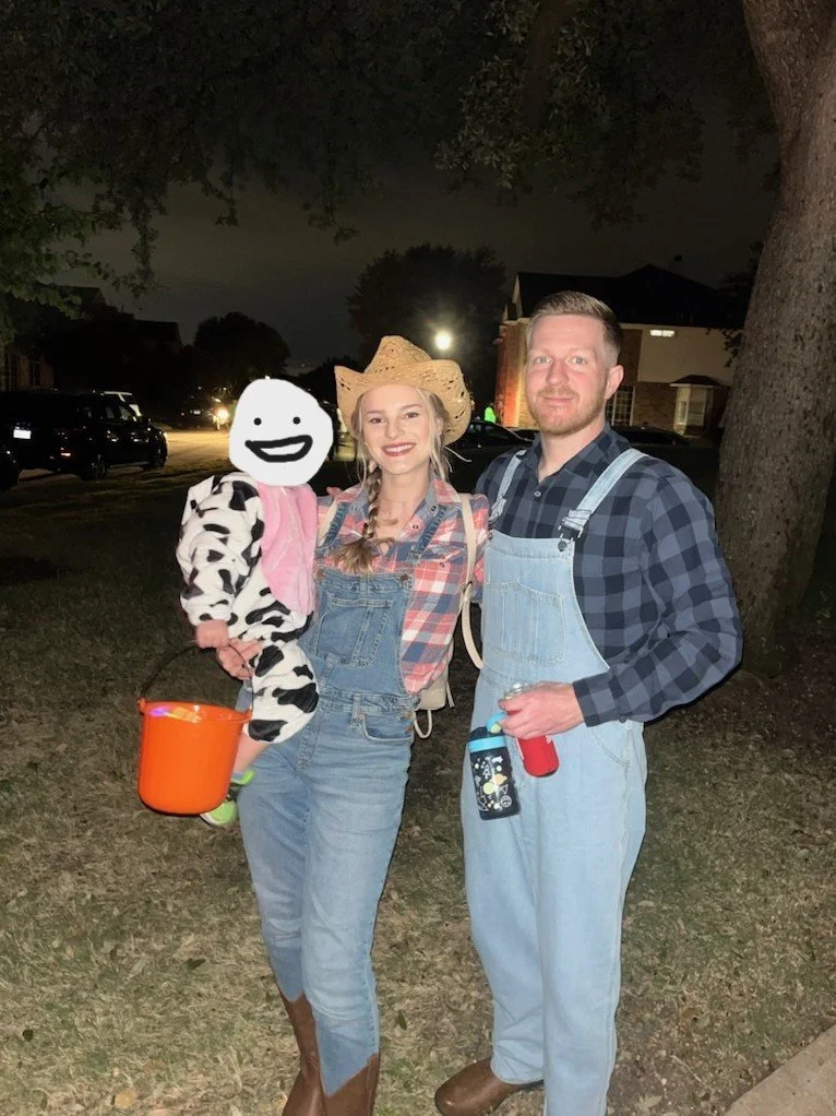 A woman dressed as a farmer with a straw hat, plaid shirt, and overalls. A man next to her wearing a checkered shirt and overalls, holding a beverage in a can. They are outdoors at night with large trees and houses in the background. A child with a doodle face mask, dressed in cow print, hold an orange pumpkin bucket.