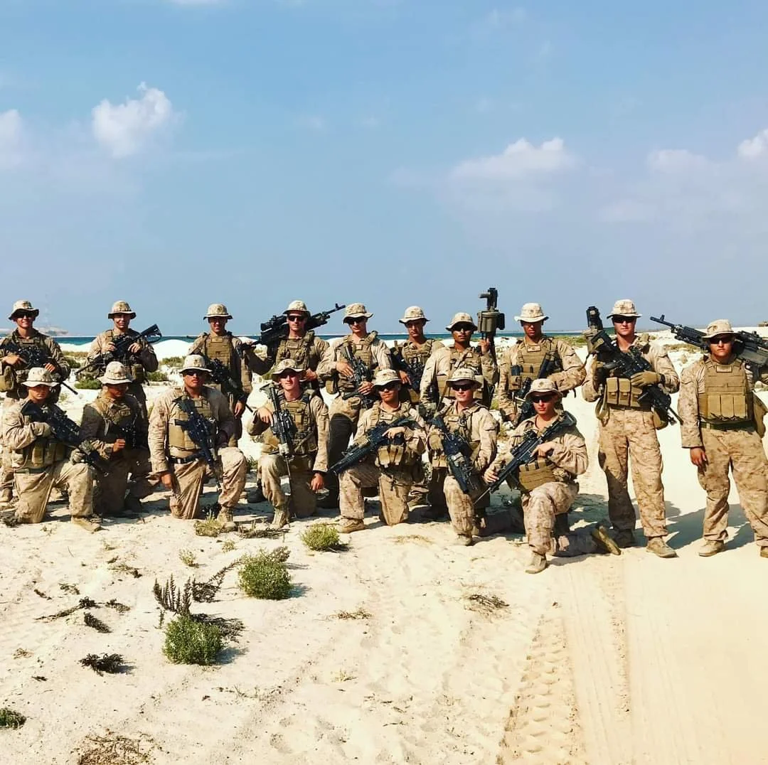 Group of Marines in desert camouflage uniforms and helmets posing on sandy terrain with a blue sky and ocean in the background, holding rifles.