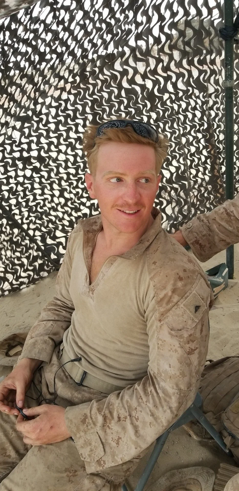 A young man with light skin and red hair, wearing desert camouflage military uniform, sunglasses on his head, sitting outdoors in front of a camouflage fabric backdrop with a sandy ground.